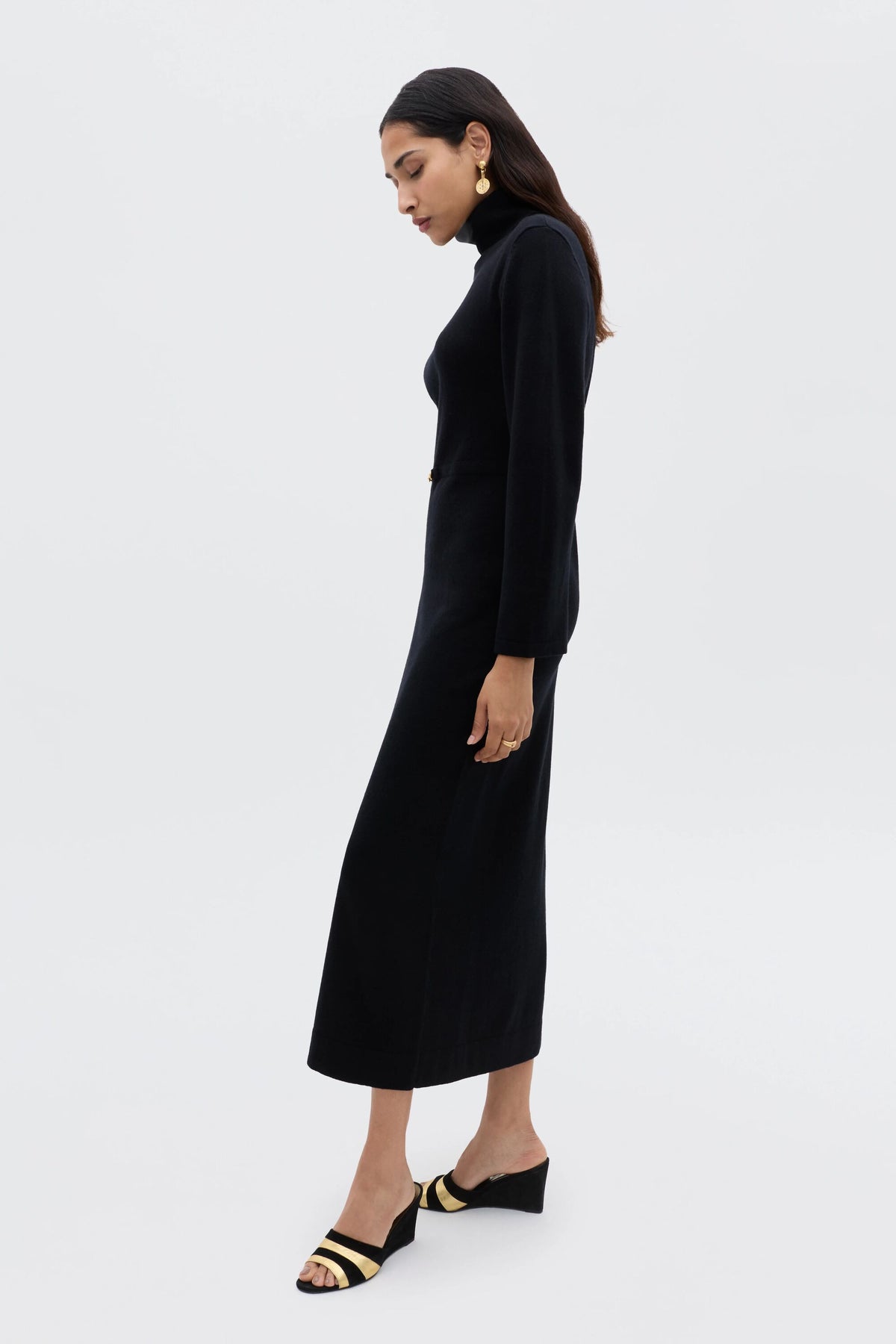 A woman stands in profile against a plain background, modeling minimalist style in the Elliette - Black long-sleeved turtleneck dress and black open-toe sandals with beige straps. She has long dark hair and gold earrings.