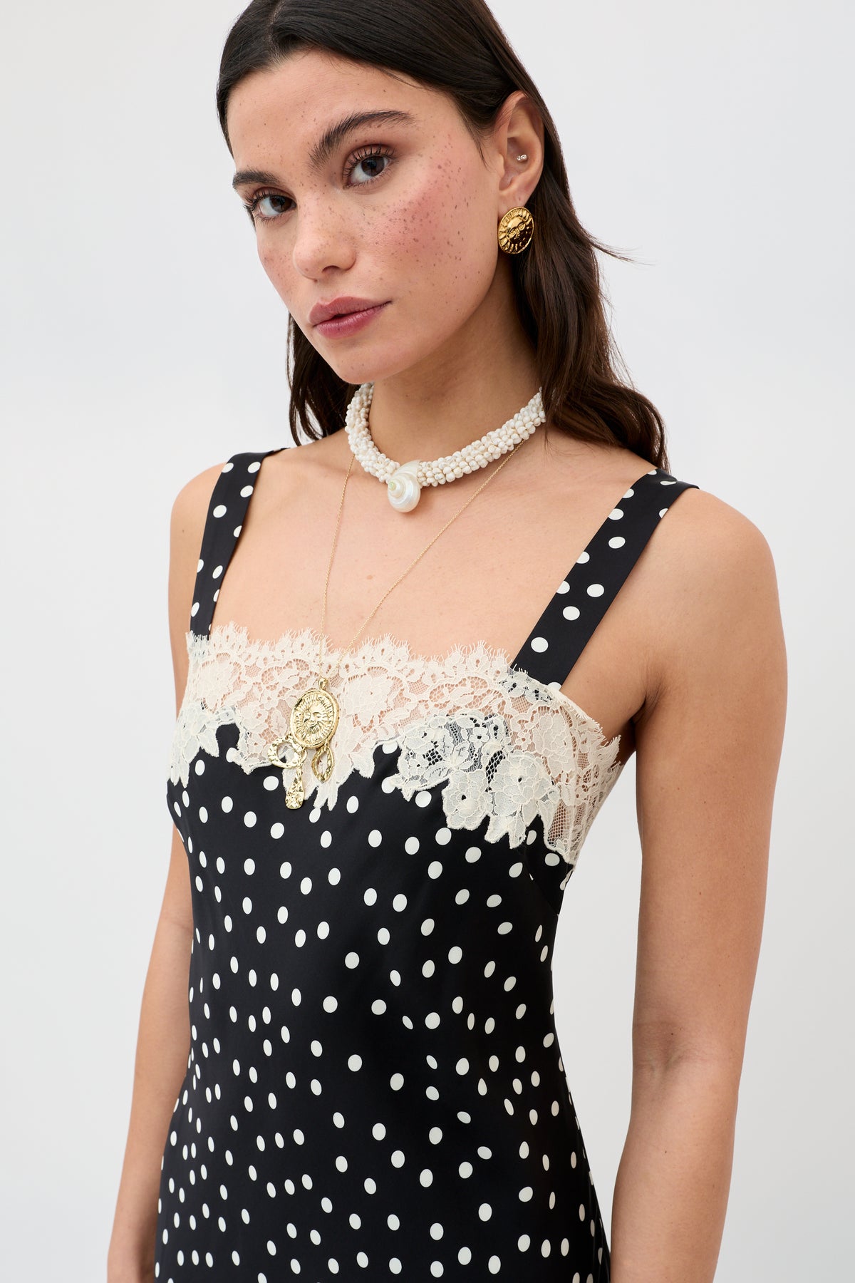 A woman wears the Noni - Gold earrings with a black polka-dot dress and lace trim, layered necklaces, and a pearl choker, standing against a plain light background.