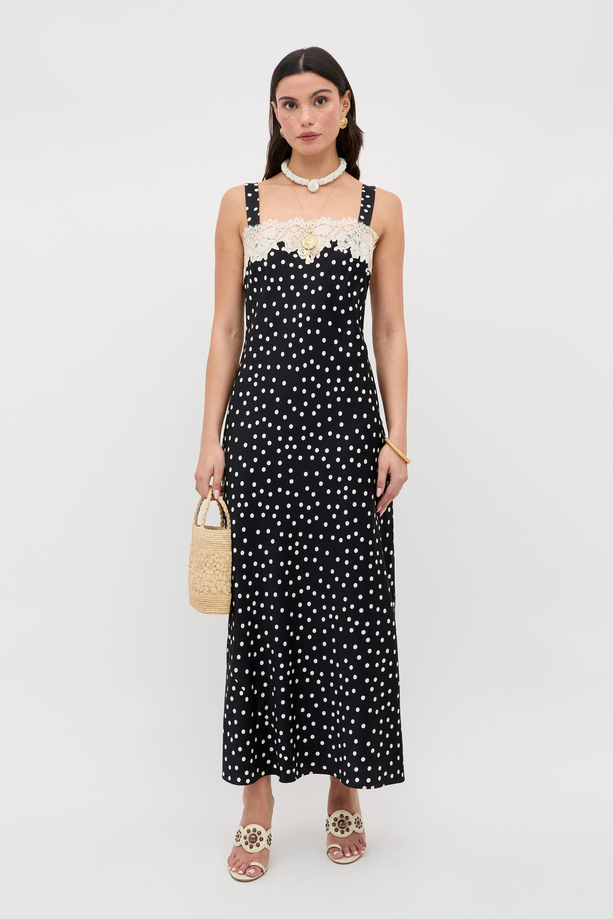 A woman stands against a plain white background wearing the Rosamarie - Scattered Dot Black dress with lace neckline, paired with beige sandals, a shell necklace, and holding a small woven handbag.