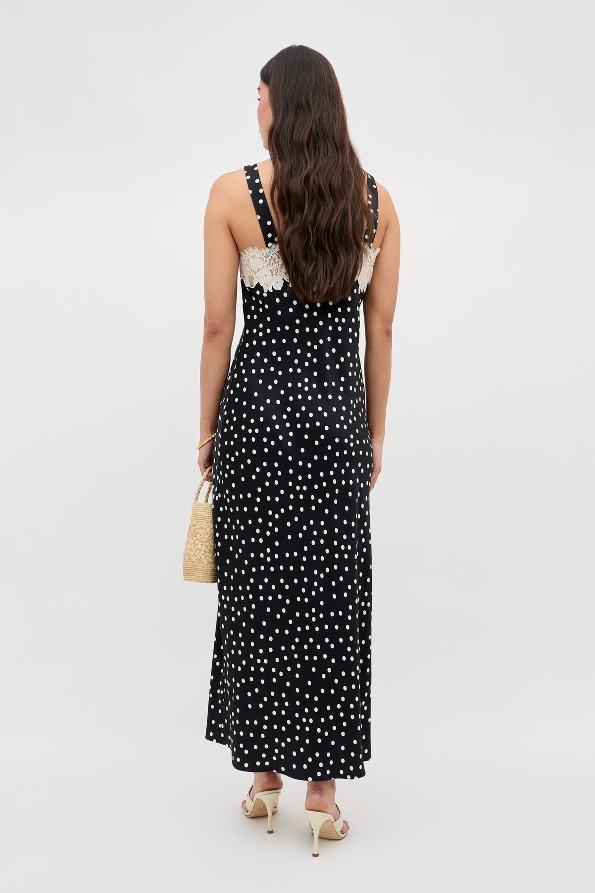 A woman with long, wavy brown hair stands facing away in the Rosamarie - Scattered Dot Black sleeveless maxi dress with white polka dots and lace trim, holding a woven handbag and wearing heeled sandals against a plain light background.