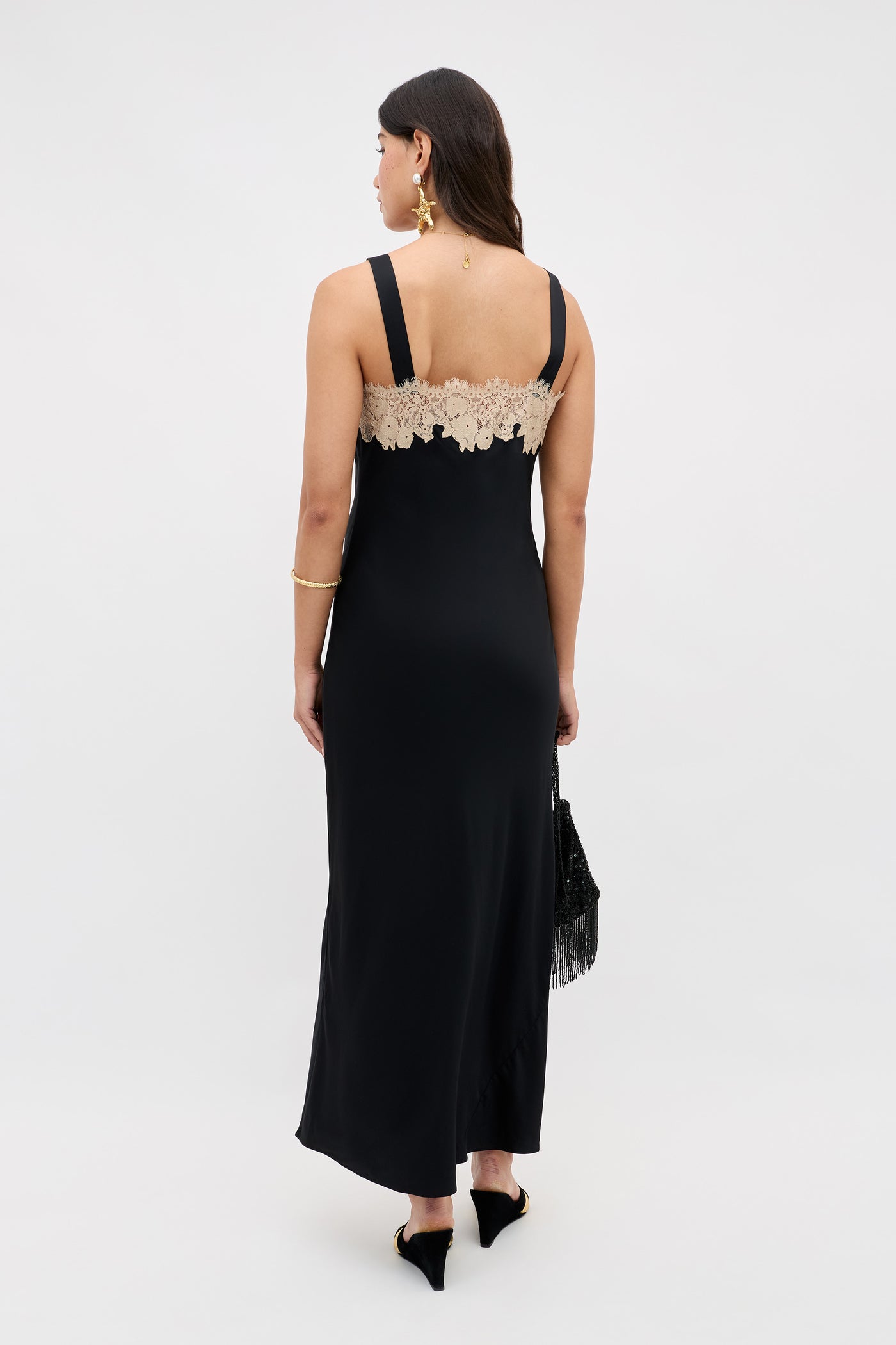 Wearing the Rosamarie - Black sleeveless gown with beige lace back, a woman faces away holding a black fringed bag. She has long dark hair down and accessorizes with gold earrings, a bracelet, and black pointed shoes.