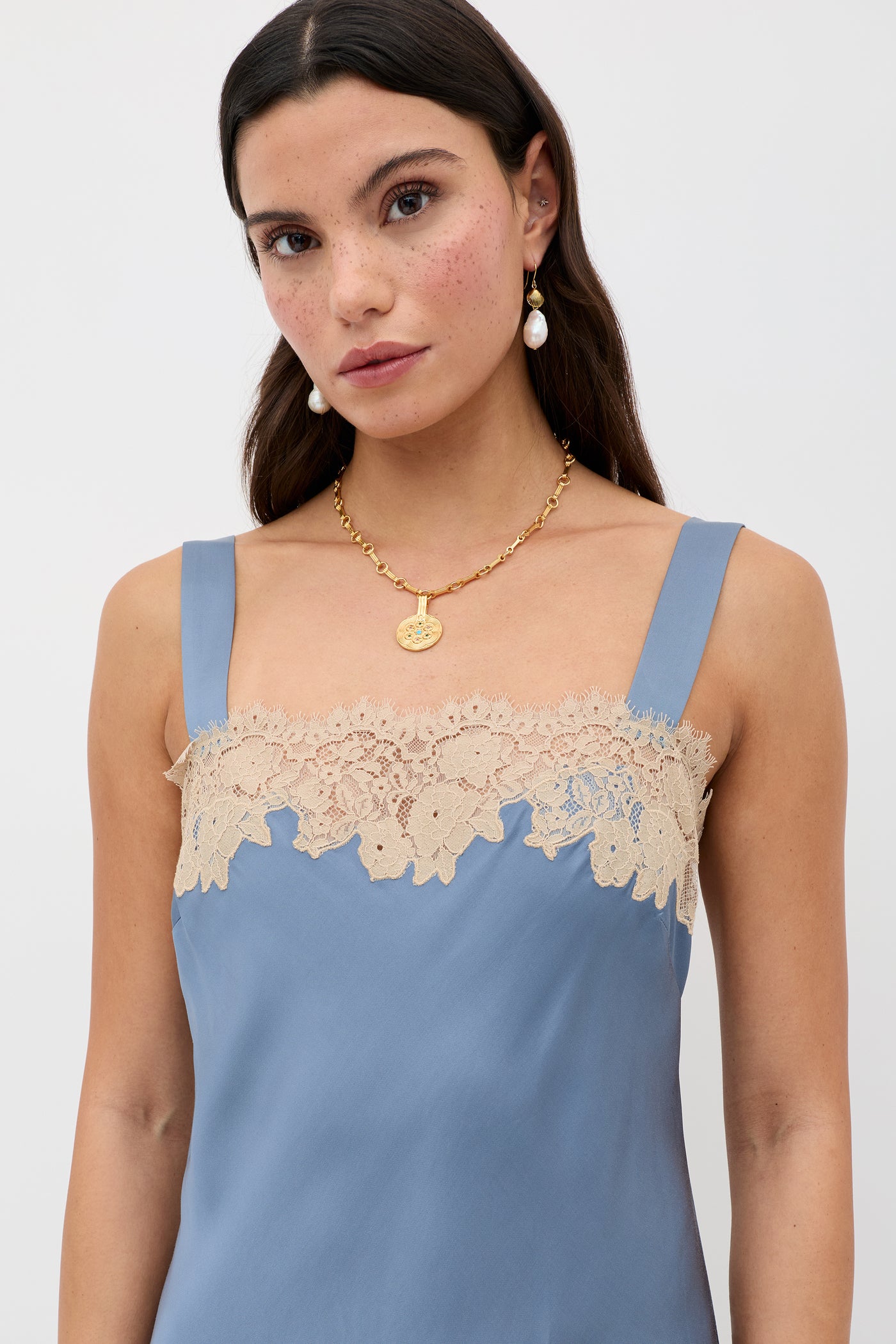 A woman with long dark hair wears the Rosamarie - Stone Blue sleeveless dress with beige lace trim, gold pendant necklace, and a pearl earring against a plain light background.