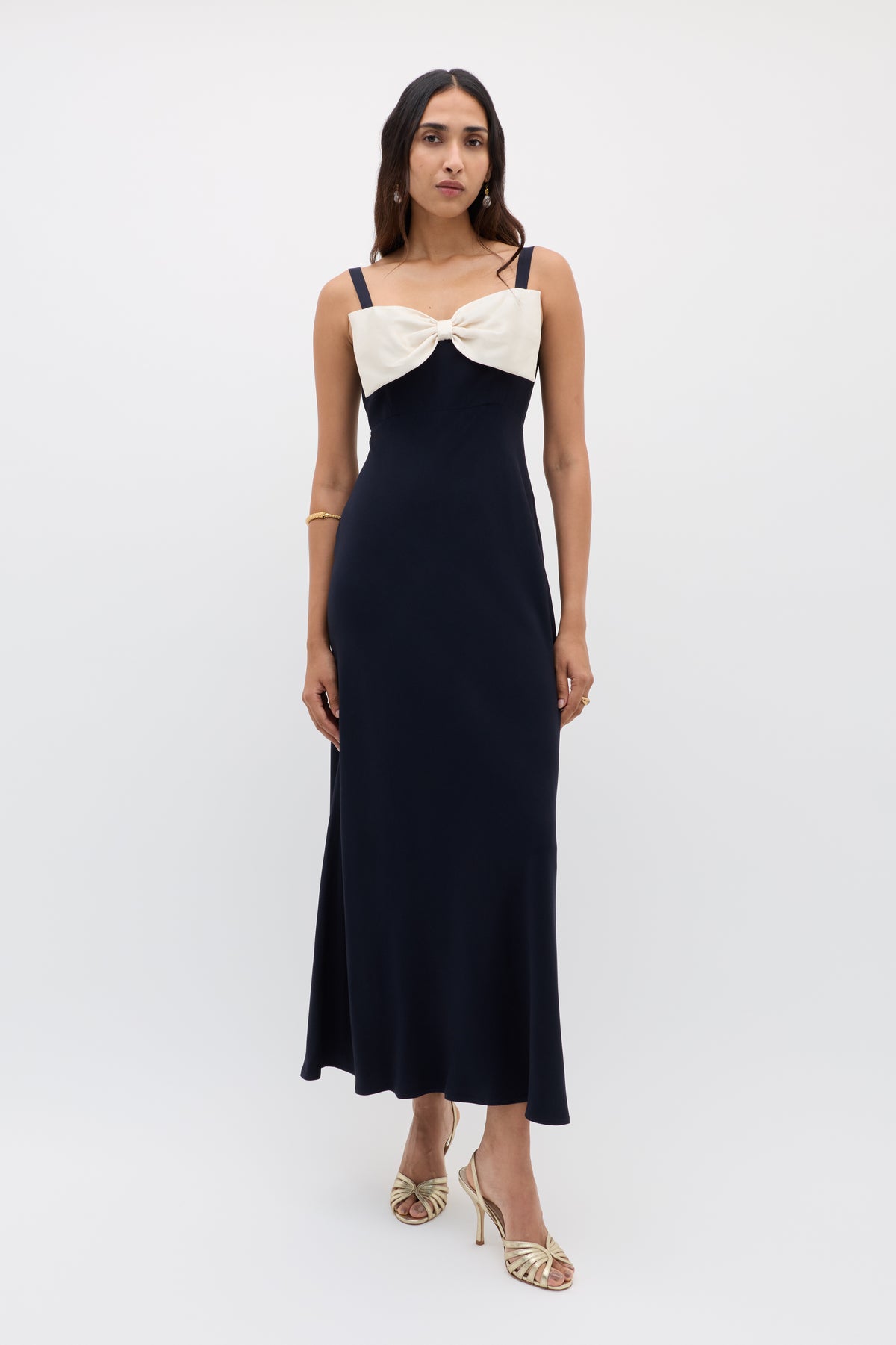 A woman wears the Melisse - Deep Navy sleeveless, floor-length evening dress with an effortless drape and a large cream bow on the chest, complemented by gold heels and a gold bracelet, against a plain white background.