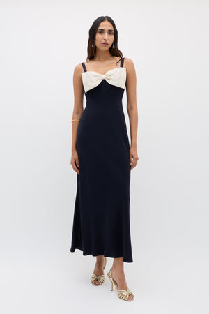 A woman wears the Melisse - Deep Navy sleeveless, floor-length evening dress with an effortless drape and a large cream bow on the chest, complemented by gold heels and a gold bracelet, against a plain white background.