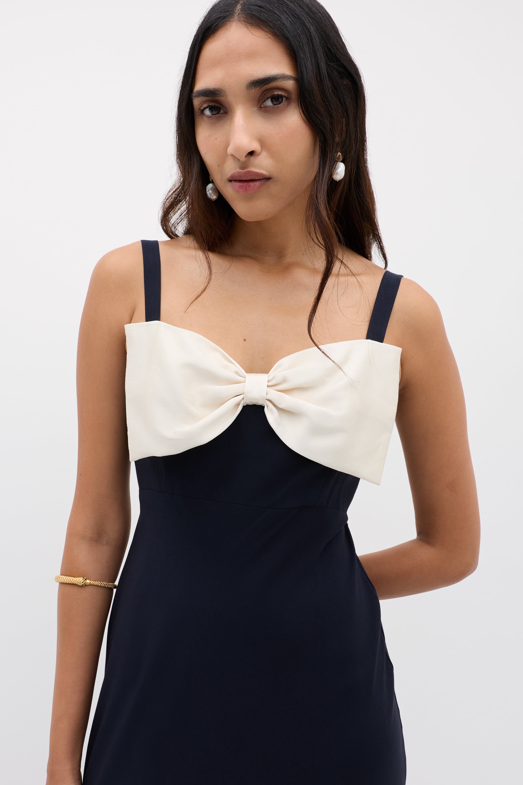 A woman in the Melisse Deep Navy sleeveless evening dress with an elegant drape and a large white bow on the chest, accessorized with pearl earrings and a gold bracelet, stands against a plain light background.