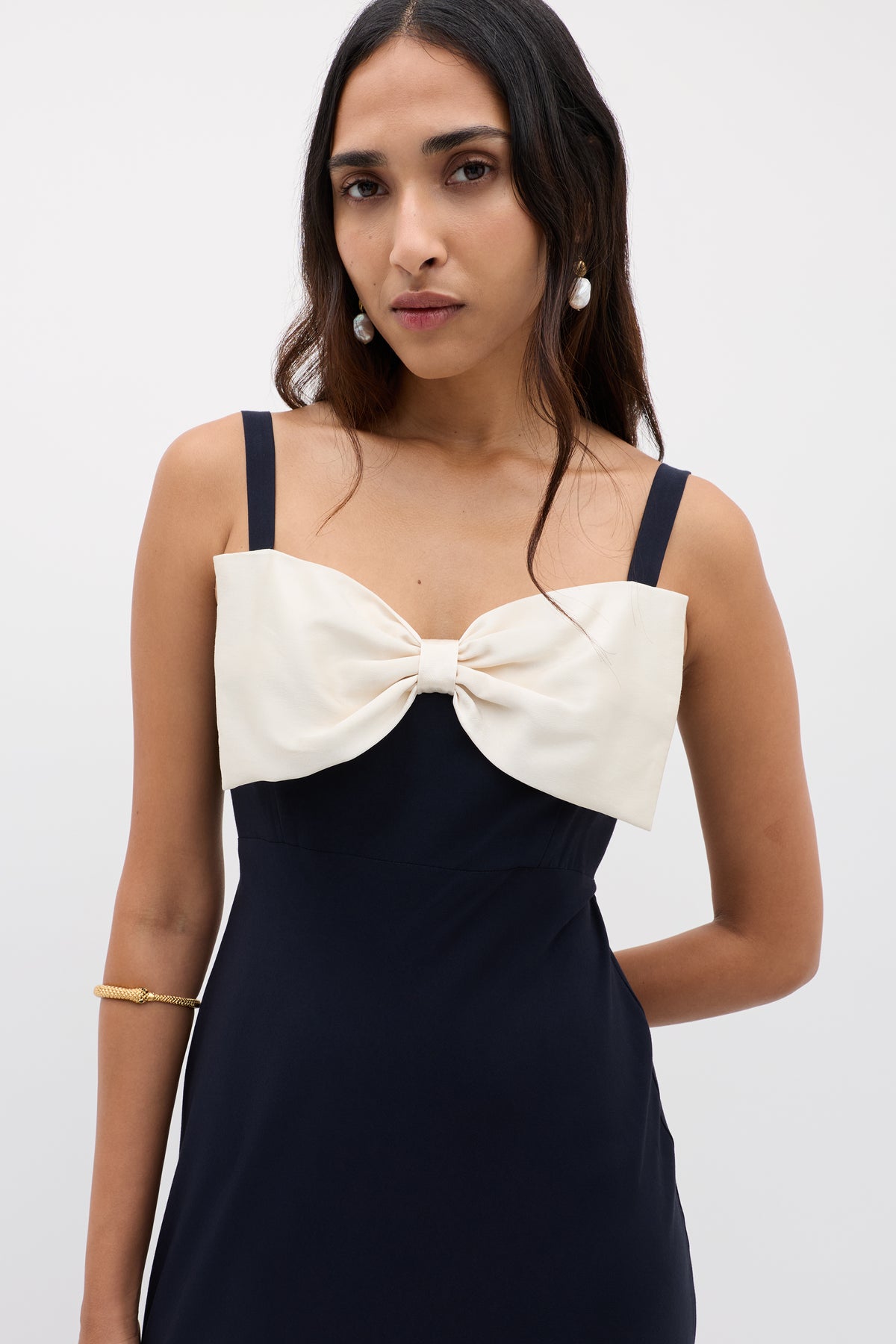 A woman in the Melisse Deep Navy sleeveless evening dress with an elegant drape and a large white bow on the chest, accessorized with pearl earrings and a gold bracelet, stands against a plain light background.