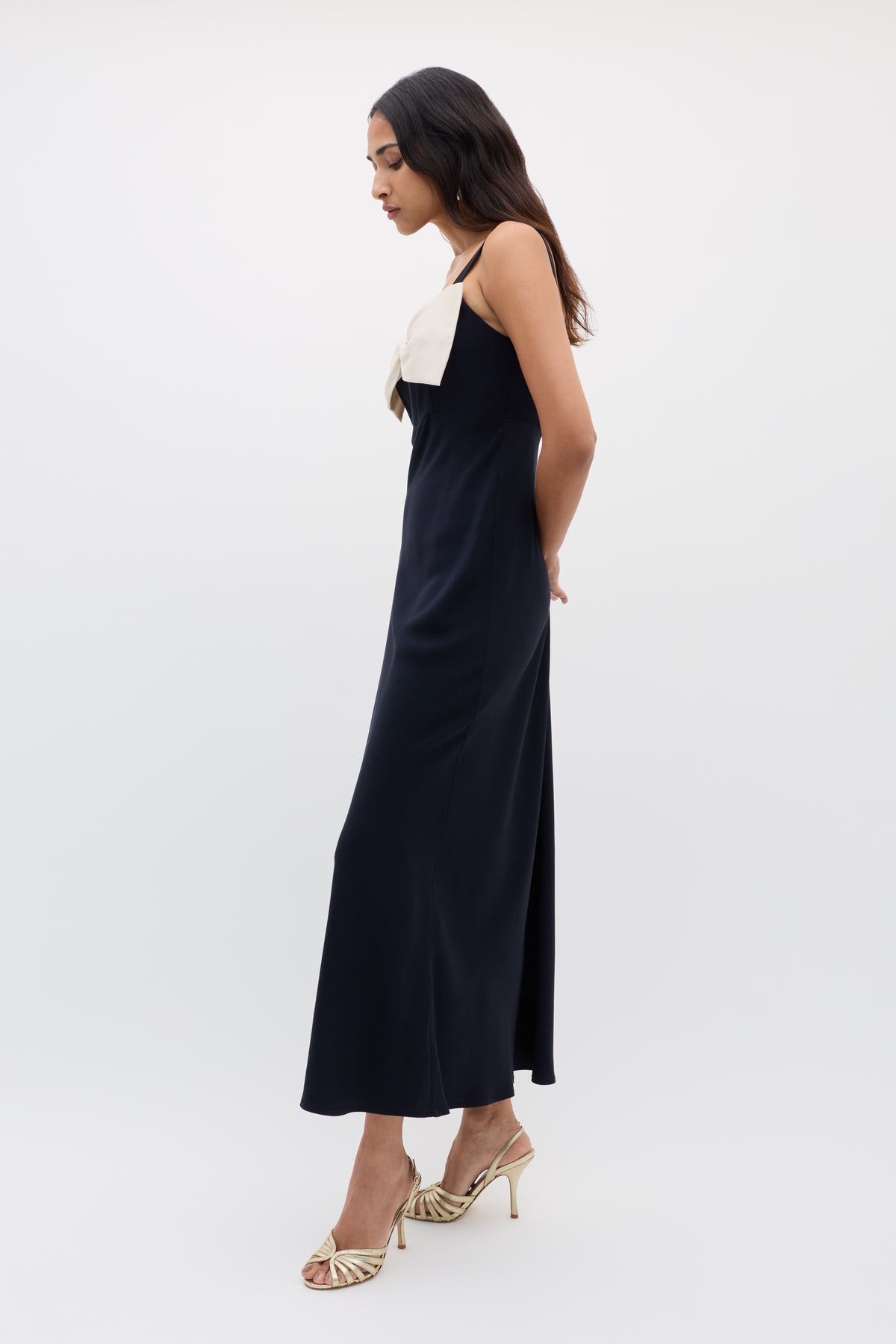 A woman stands in profile against a white background, wearing the Melisse - Deep Navy sleeveless, ankle-length evening dress with an effortless drape, a large bow detail on the chest, and gold high-heeled sandals.