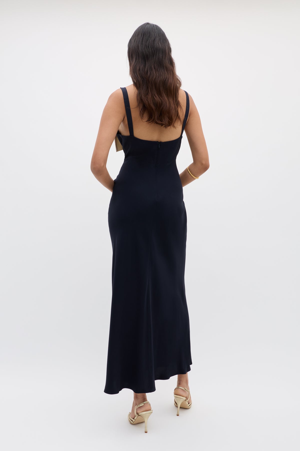 A woman with wavy brown hair stands with her back to the camera against a plain light background, wearing the Melisse - Deep Navy long, sleeveless evening dress with an effortless drape and beige high-heeled sandals.