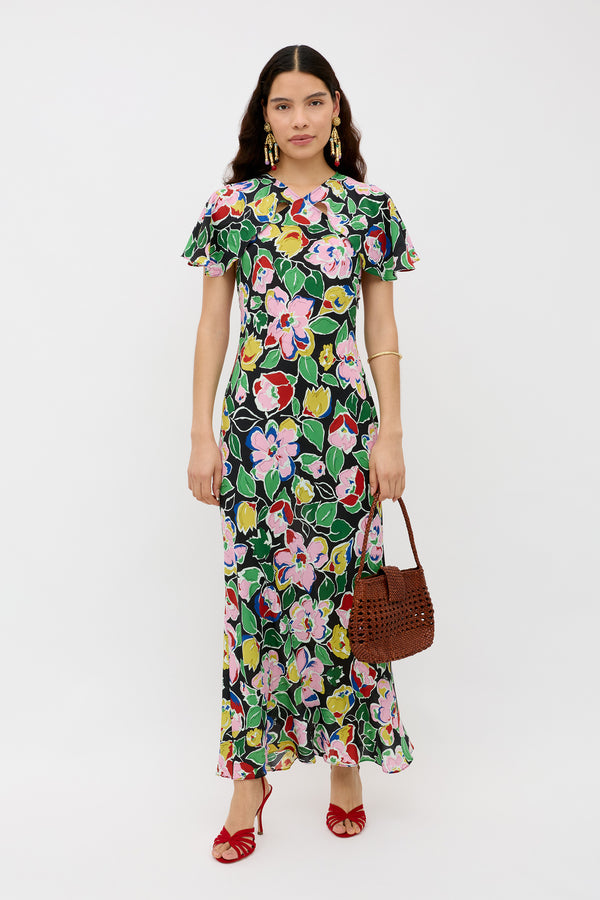 A woman stands against a white background wearing the Aryella Jeannie Bloom Black floral maxi dress with short ruffled sleeves, red strappy heels, gold earrings, a gold bangle, and holding a brown woven handbag.