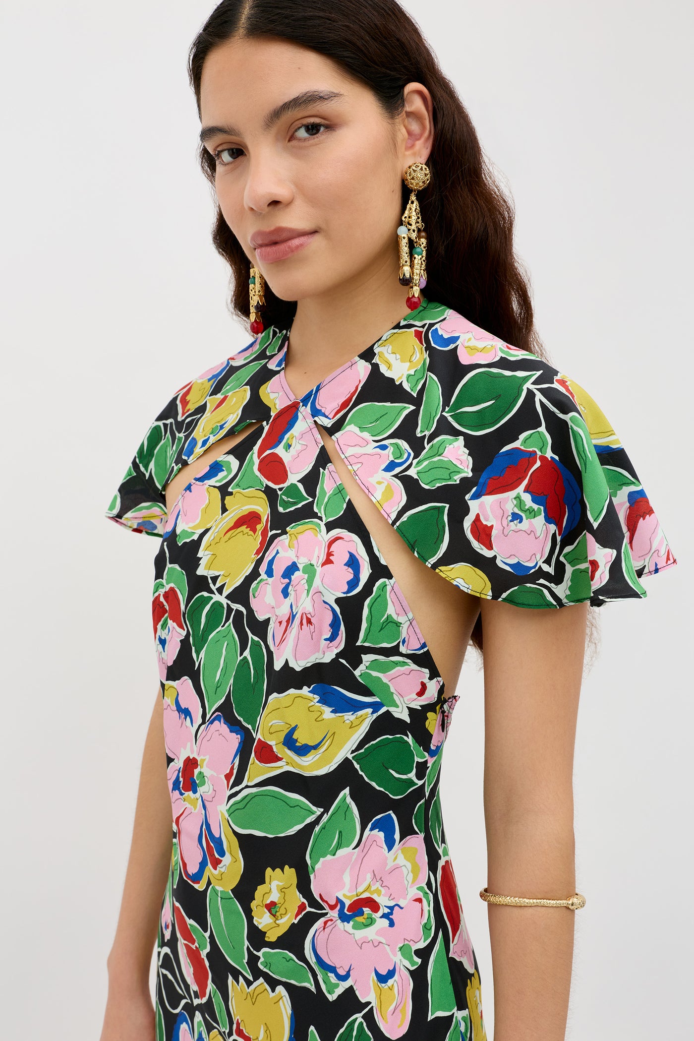 A woman with long dark hair wears the Aryella Jeannie Bloom Black dress, featuring colorful florals and cape sleeves. She pairs it with large earrings and stands against a plain light background, looking at the camera with a slight smile.