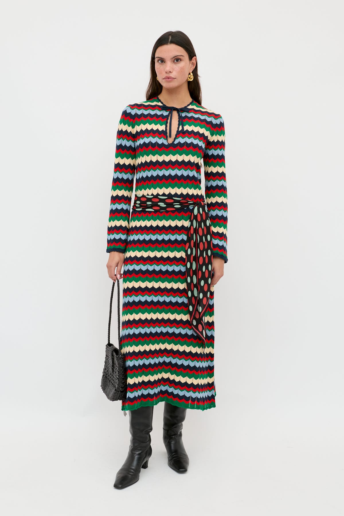 A woman is wearing the Lucila - Retro Chevron Blue long-sleeved dress, featuring a multicolored wavy chevron pattern and a keyhole neckline, styled with a patterned belt, black boots, and a black textured handbag, standing against a plain white background.