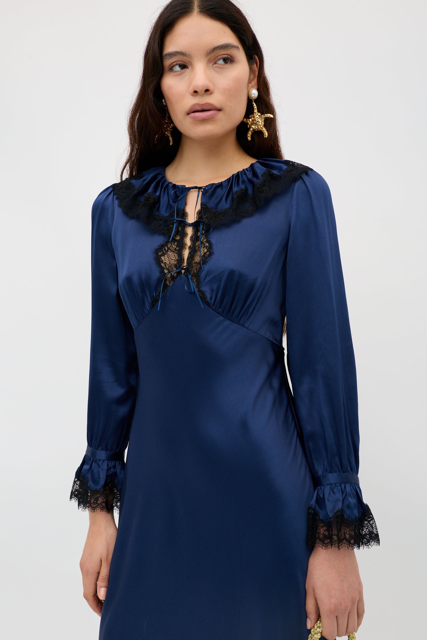 A woman with long dark hair and statement earrings wears the Brecia - Midnight Navy, a long-sleeved satin dress with black lace trim on the neckline, cuffs, and chest, standing against a plain light background.