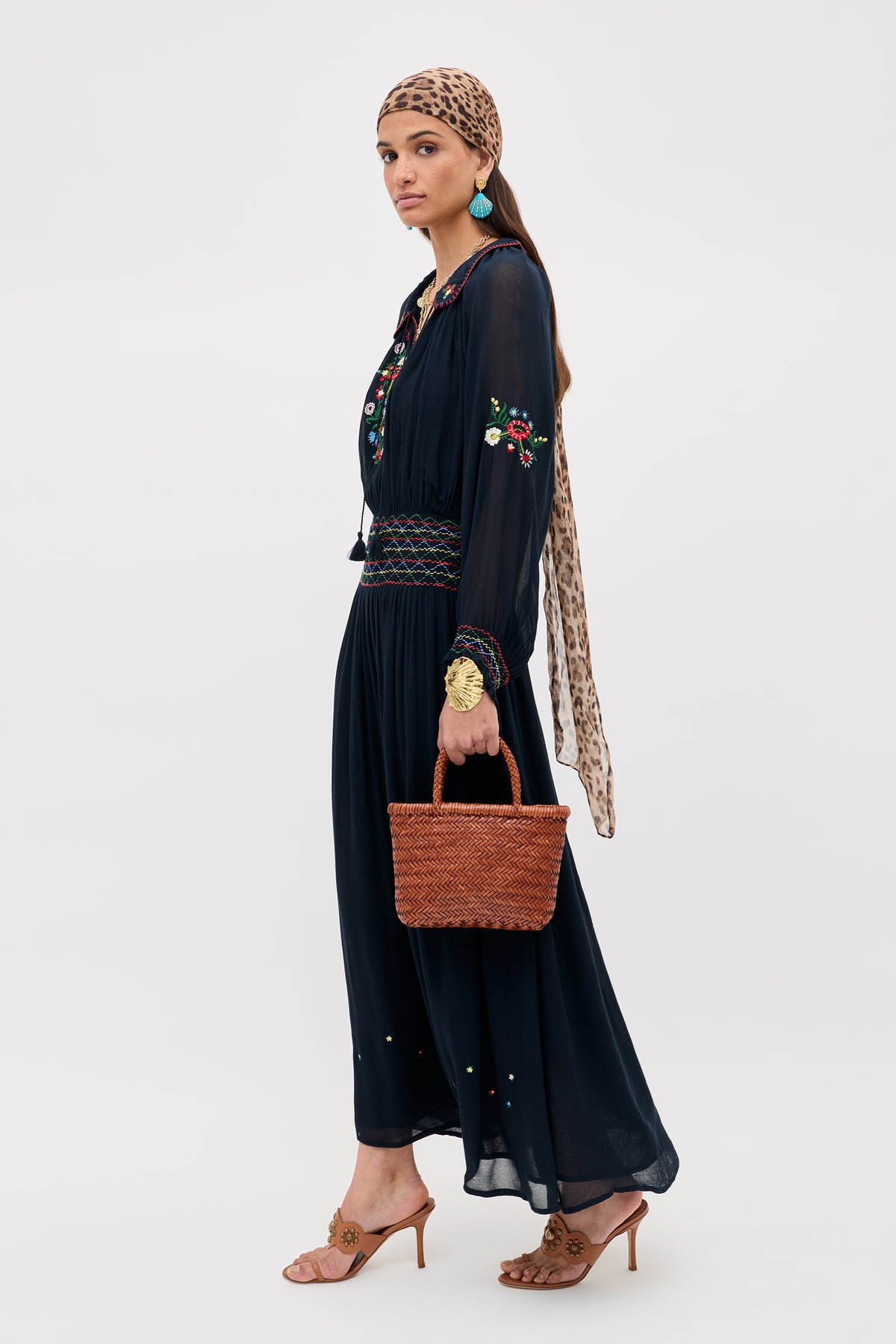 A woman stands against a plain light background wearing the Emilio - Petunia Embroidery Navy dress, tan heeled sandals, turquoise earrings, a leopard print headscarf, and holding a woven brown handbag.