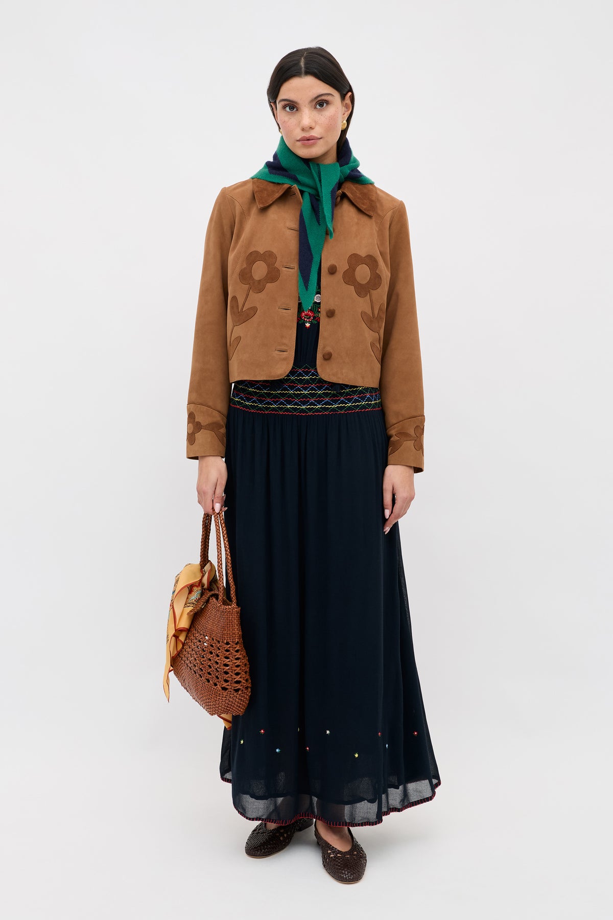 A woman faces forward in the Emilio - Petunia Embroidery Navy jacket, a green scarf, long black skirt, brown woven shoes, and carries a brown woven bag with a yellow scarf attached.