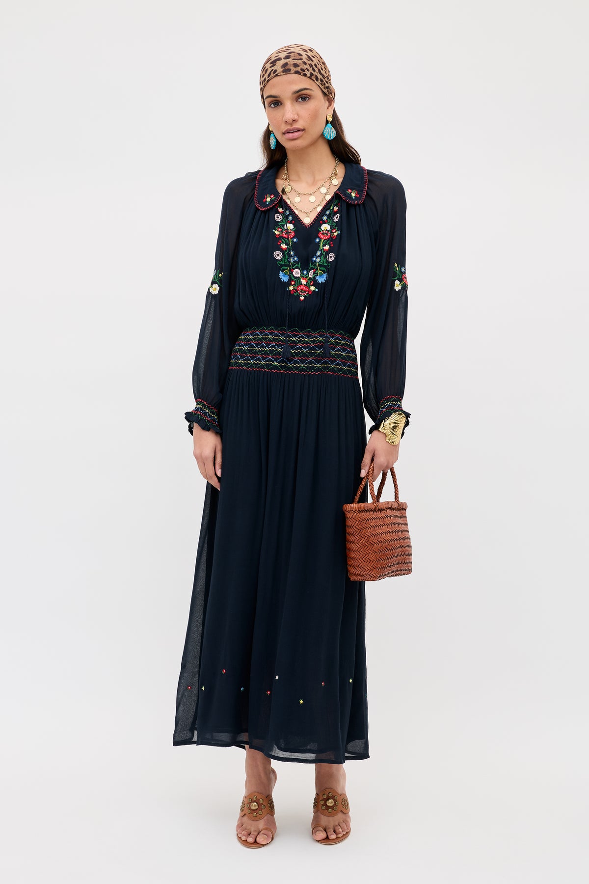 A woman wears the Emilio - Petunia Embroidery Navy long dress with long sleeves, brown sandals, a woven handbag, layered necklaces, a patterned headscarf, and hoop earrings against a plain background.