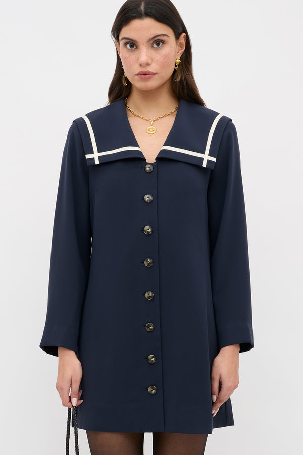 A woman models the Karina - Navy dress, featuring a white-trimmed sailor collar and black front buttons, as she stands against a plain white background holding a black bag in her left hand.