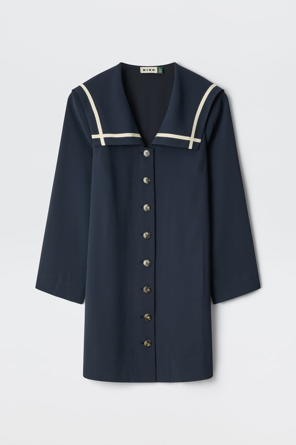 The Karina - Navy is a navy blue long-sleeve dress with a sailor collar, white trim, and front button closure. Displayed against a plain white background.