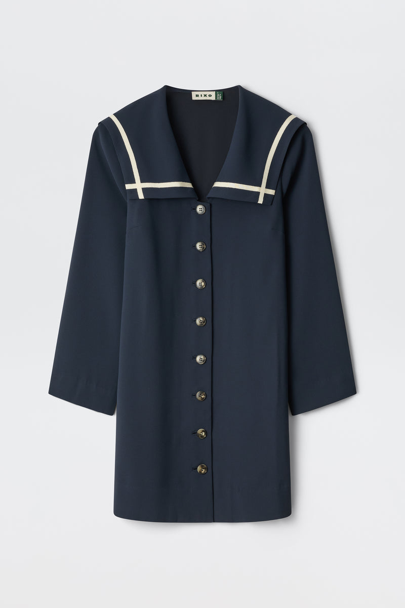 The Karina - Navy is a navy blue long-sleeve dress with a sailor collar, white trim, and front button closure. Displayed against a plain white background.