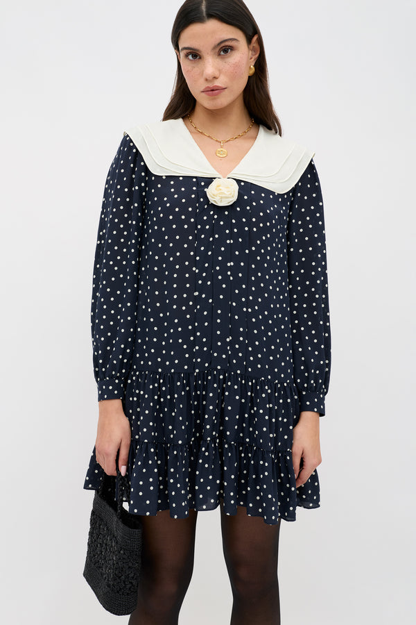 A woman in the Valetta Layla Dot Navy dress—navy blue with white polka dots, a sailor collar, long sleeves, ruffled hem, and a decorative flower—holds a small black woven bag.