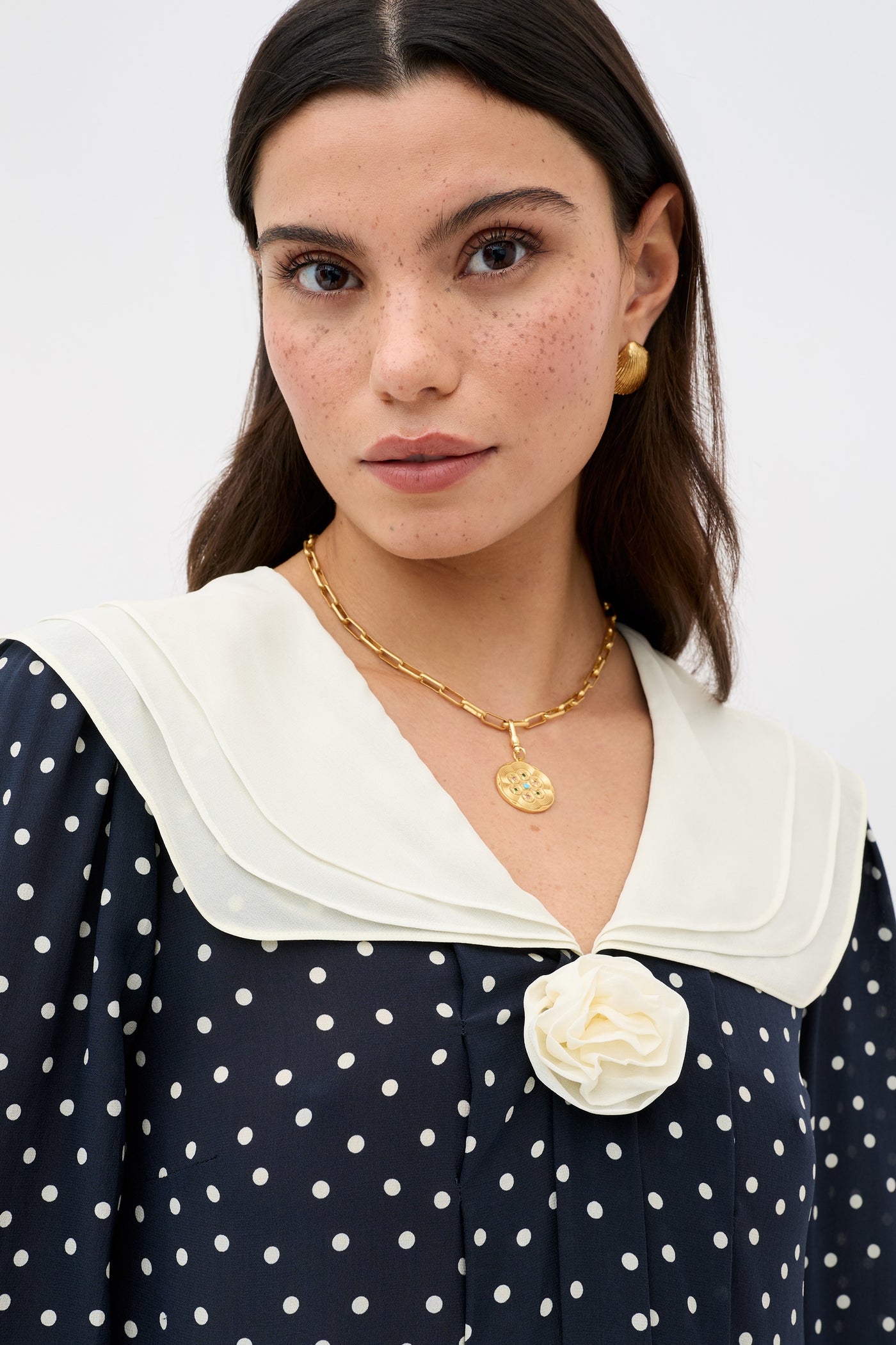 A woman with long brown hair and freckles wears the Valetta Layla Dot Navy dress, featuring white polka dots, a white collar, a gold necklace with round pendant, gold earrings, and a white flower detail at the neckline.