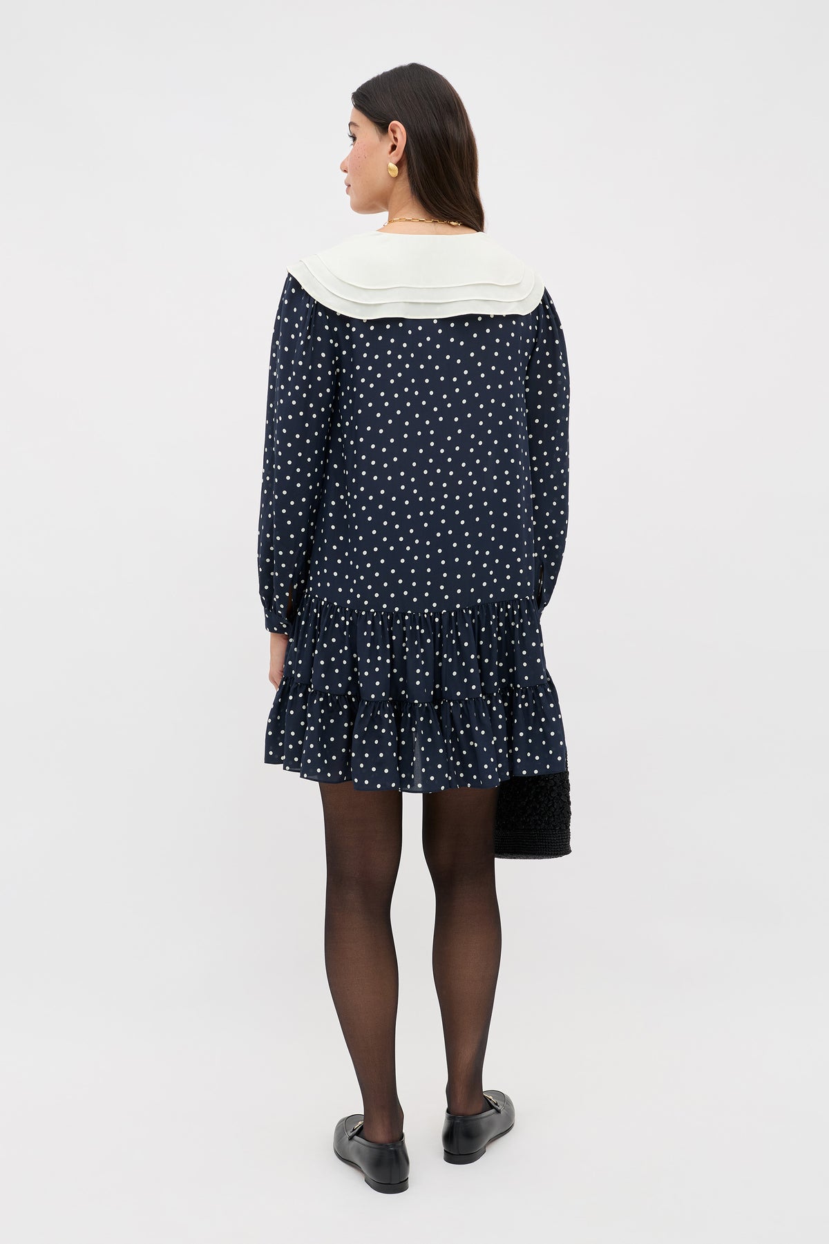 A woman with long brown hair faces away, wearing the Valetta - Layla Dot Navy dress featuring white polka dots and a white collar. She pairs it with black tights, shoes, and a black textured bag against a plain light background.
