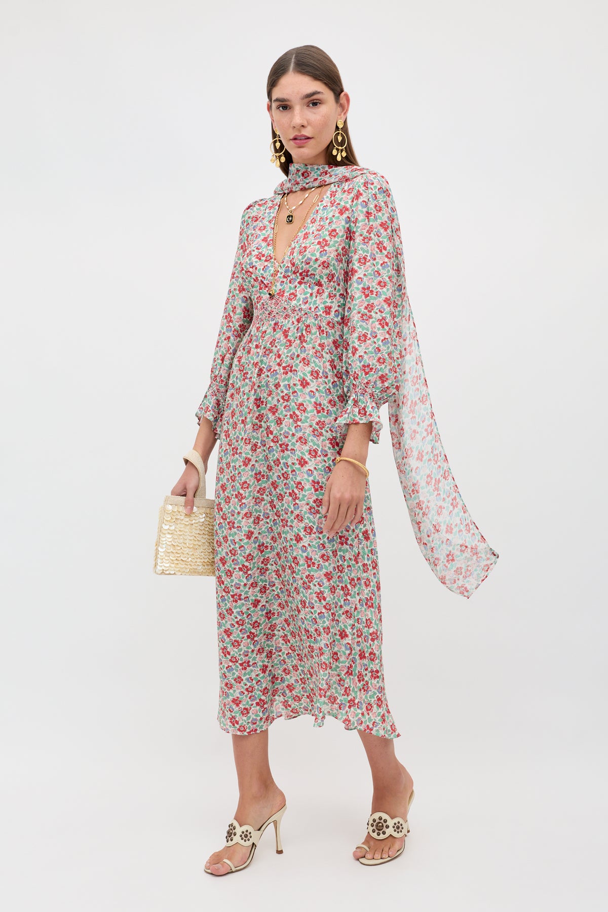 A woman models the Mireta - Jeannie Bloom Ivory, a long floral dress with a deep V-neck and flowing sleeves, paired with gold earrings, layered necklaces, a woven clutch, and beige heeled sandals against a plain white background.