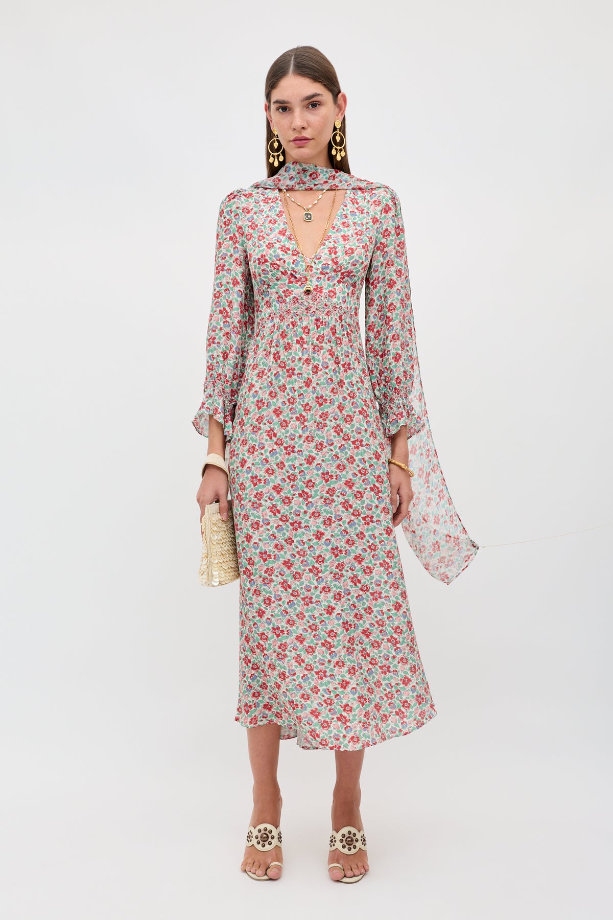 A woman stands against a plain light background wearing the Mireta - Jeannie Bloom Ivory long, floral print dress with flared sleeves, styled with gold earrings, a choker necklace, white heeled sandals, and a woven clutch.