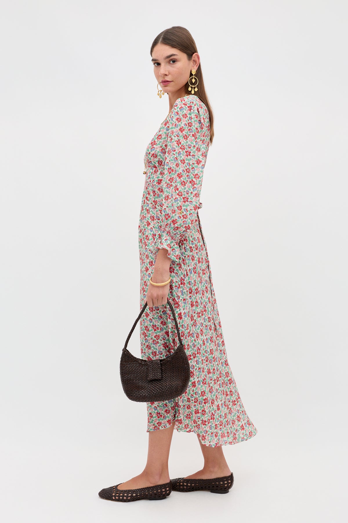 A woman stands sideways against a plain background, wearing the Mireta - Jeannie Bloom Ivory long floral dress, large gold earrings, brown woven flats, and holding a dark brown textured handbag as she looks toward the camera.
