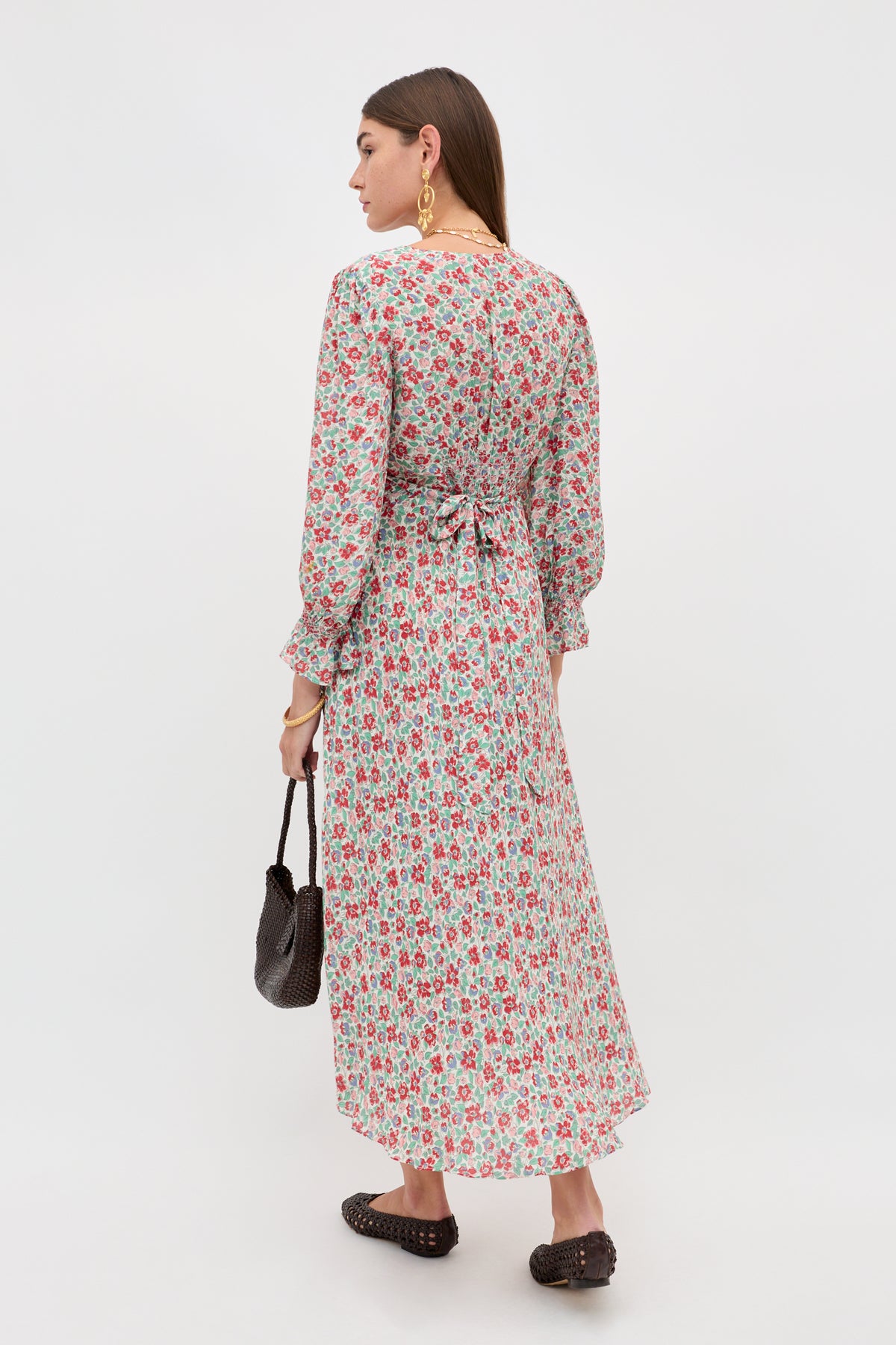 A woman stands with her back turned, wearing the Mireta - Jeannie Bloom Ivory long-sleeved, ankle-length floral dress with a waist tie. She carries a small black handbag, wears black slip-on shoes, and large gold earrings.