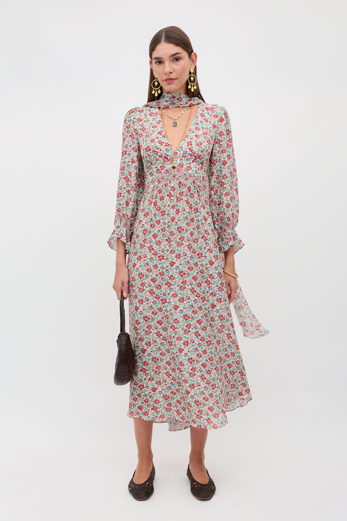 A woman poses against a white background in the Mireta - Jeannie Bloom Ivory, a long-sleeved, floral print midi dress with a V-neckline. She completes her look with gold earrings, layered necklaces, a brown woven bag, and black flats.