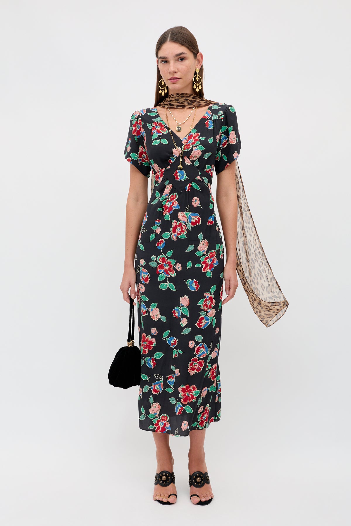 A woman stands against a white background wearing the Riella - Kimmie Floral Charcoal dress, black sandals, statement earrings, a sheer leopard-print scarf, and holding a small black handbag.