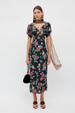 A woman stands against a white background wearing the Riella - Kimmie Floral Charcoal dress, black sandals, statement earrings, a sheer leopard-print scarf, and holding a small black handbag.