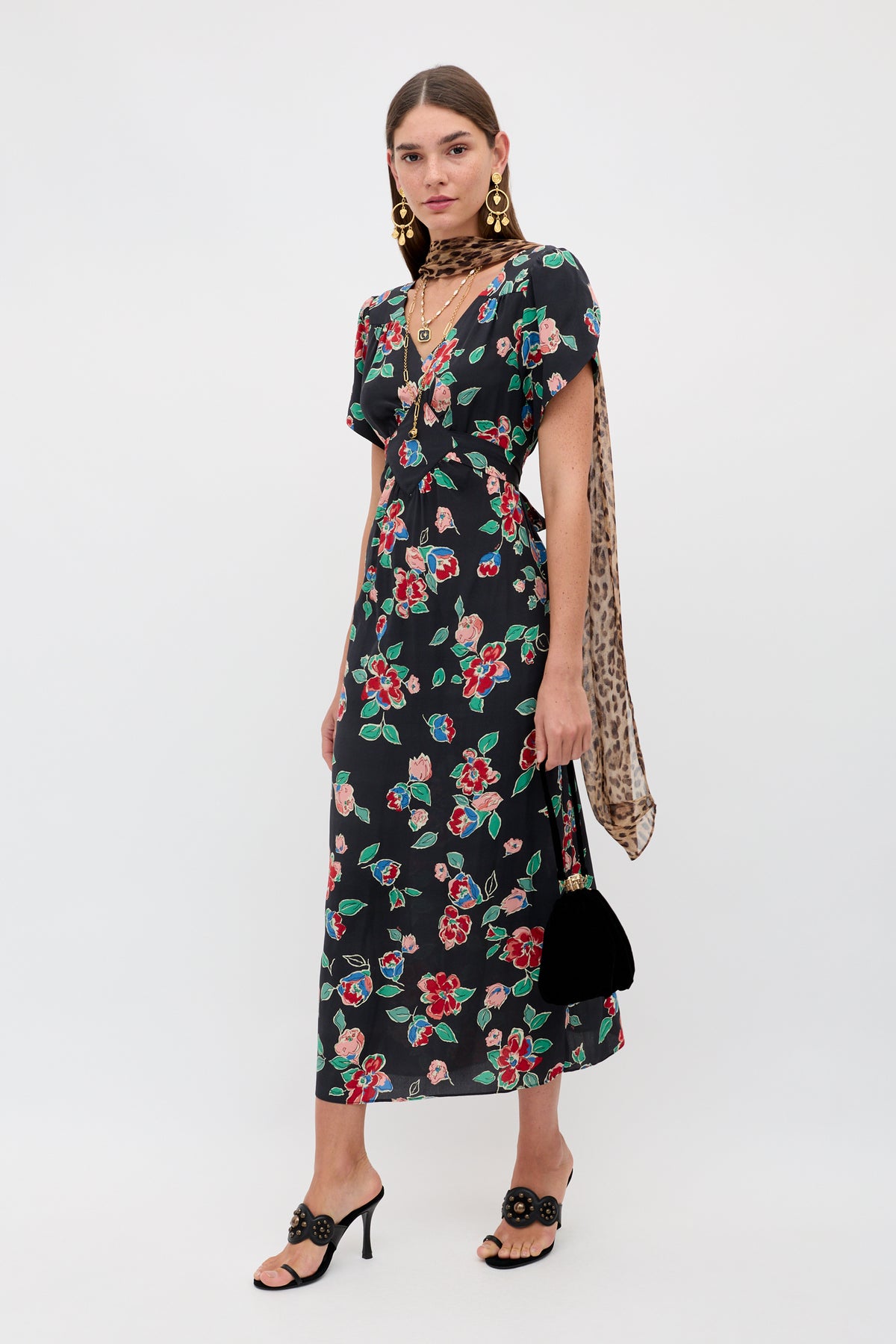 A woman wearing the Riella - Kimmie Floral Charcoal dress, black heeled sandals, gold earrings, layered necklaces, a leopard print scarf, and holding a black handbag poses against a plain white background.
