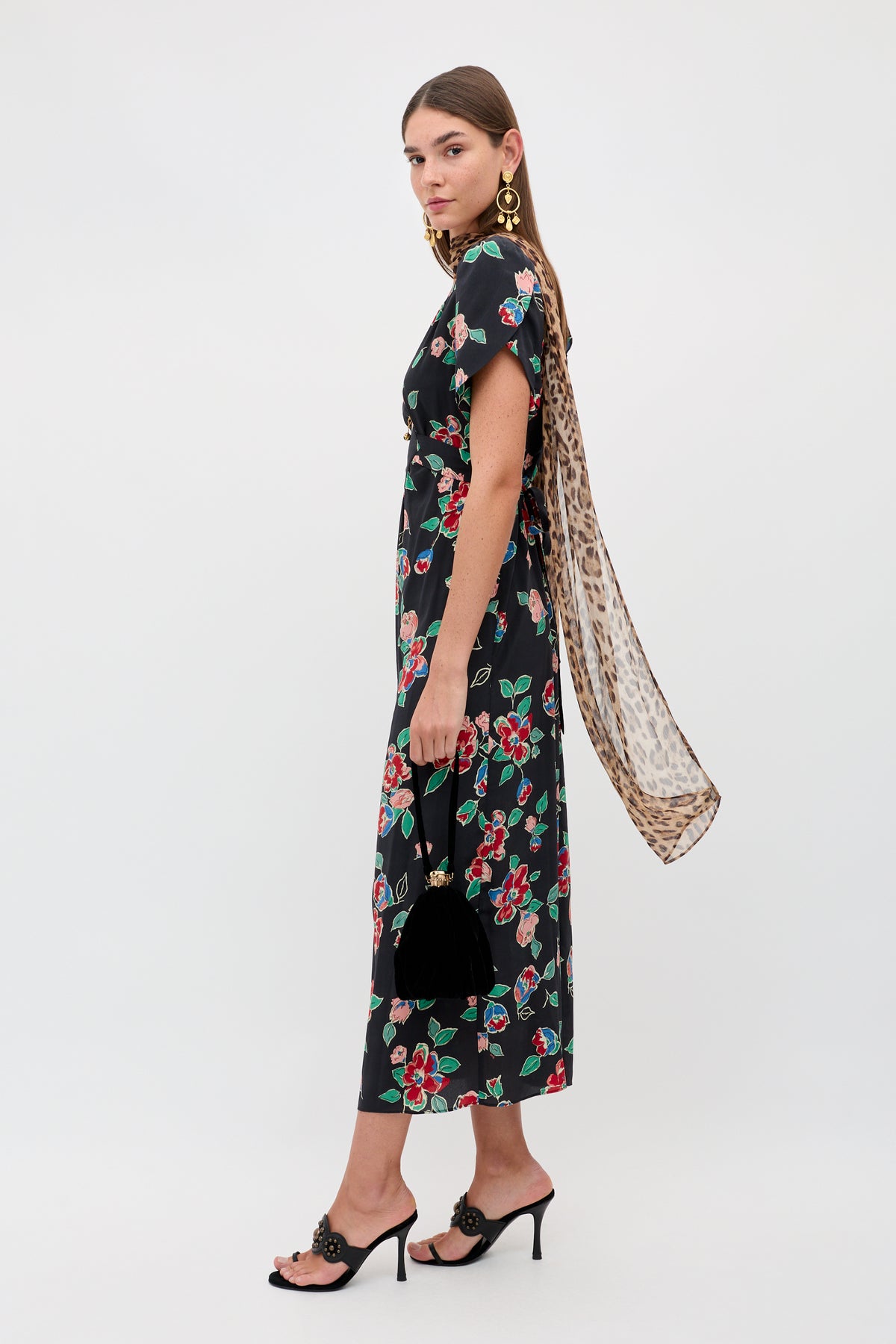 A woman stands sideways in the Riella - Kimmie Floral Charcoal midi dress with black high-heeled sandals, statement gold earrings, a leopard-print scarf, and a black handbag, posing against a plain white background.