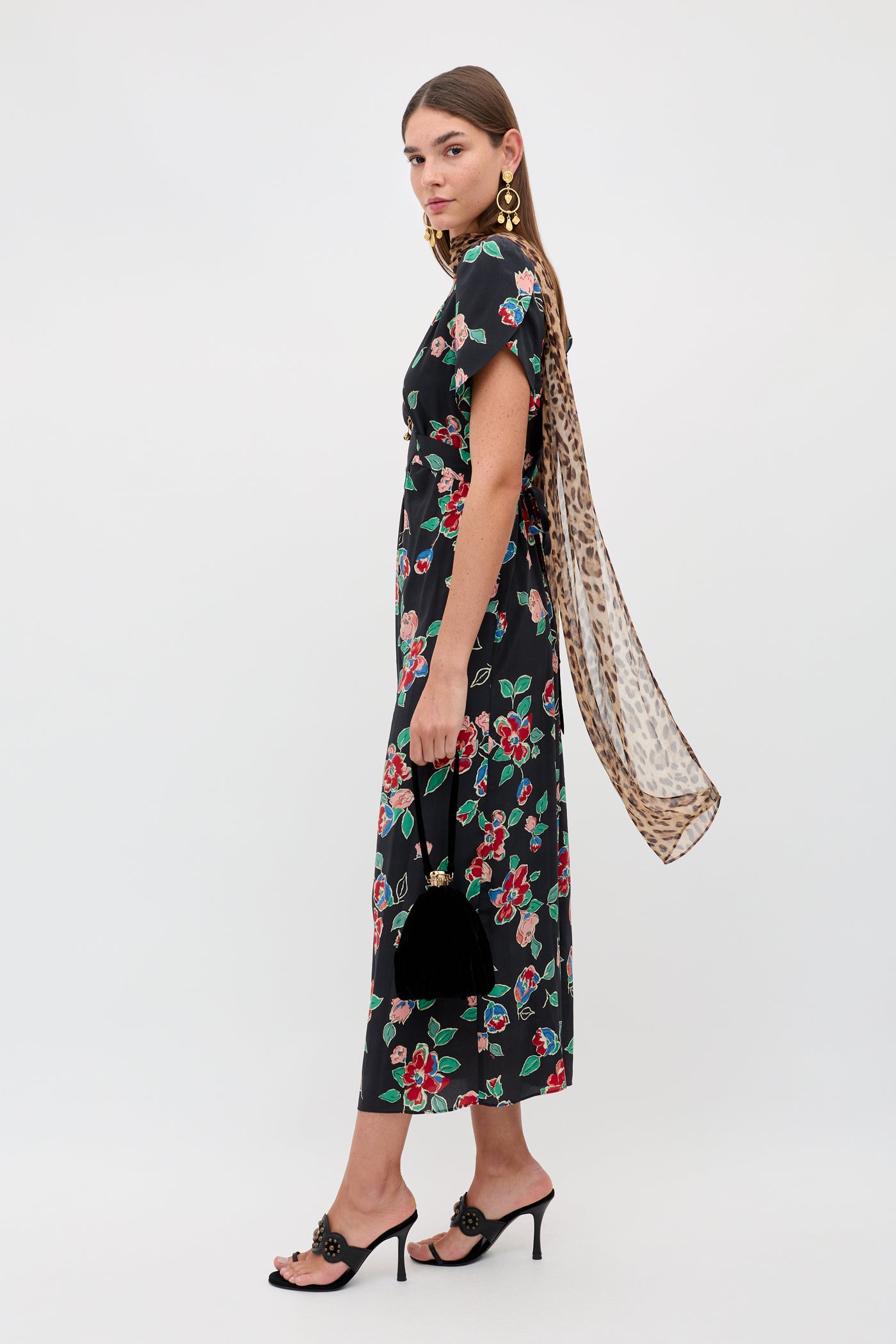 A woman stands sideways in the Riella - Kimmie Floral Charcoal midi dress with black high-heeled sandals, statement gold earrings, a leopard-print scarf, and a black handbag, posing against a plain white background.