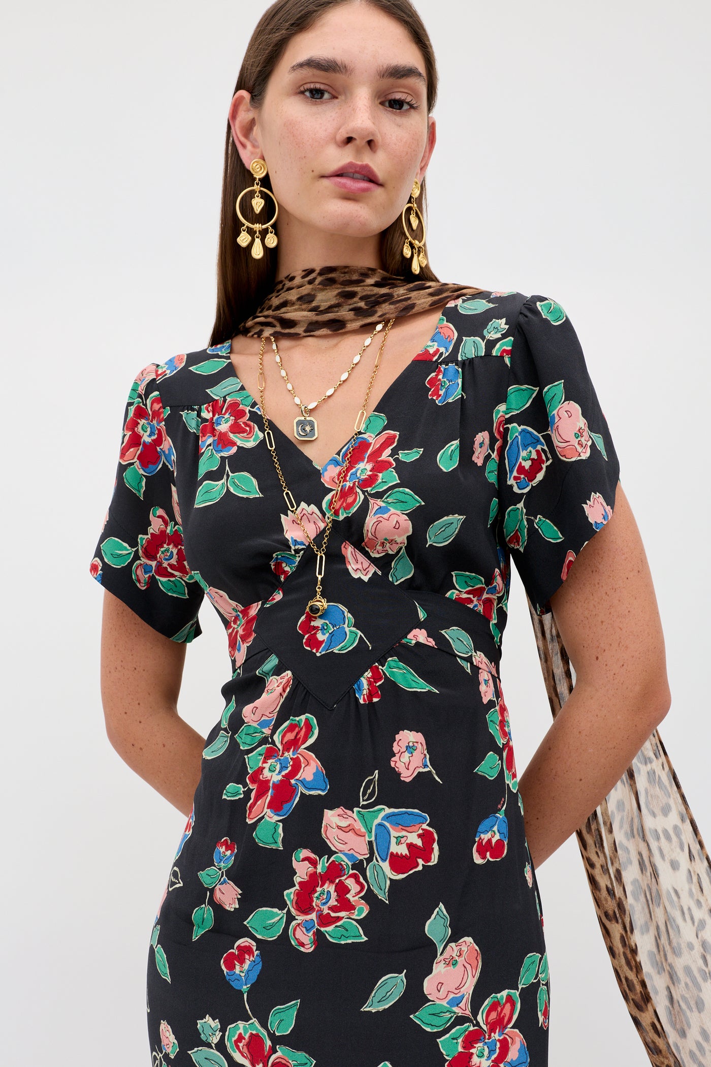 A woman wearing the Riella - Kimmie Floral Charcoal dress, gold earrings, layered gold necklaces, and a leopard print scarf poses against a plain light background.