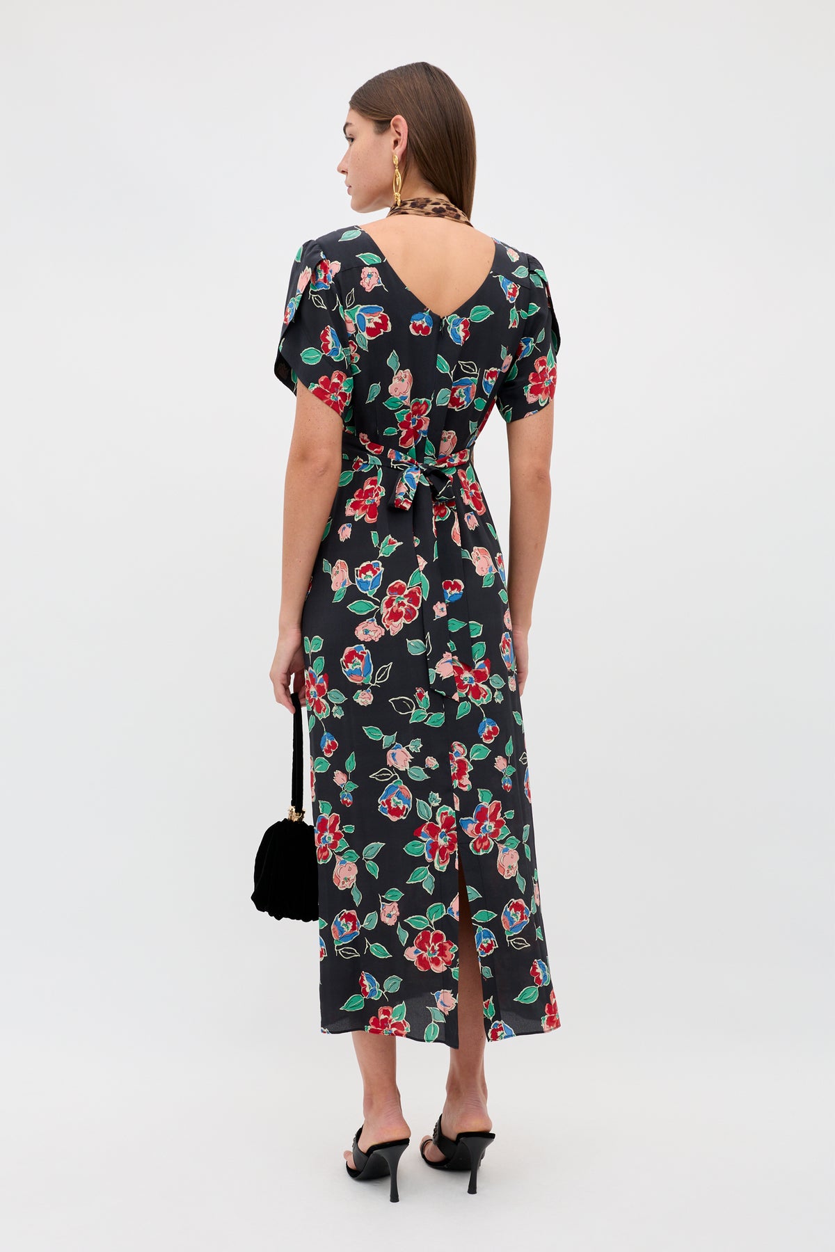 A woman faces away wearing the Riella - Kimmie Floral Charcoal ankle-length dress with short sleeves and a belt, paired with black heeled sandals, a small black handbag, large earrings, and her long brown hair down.