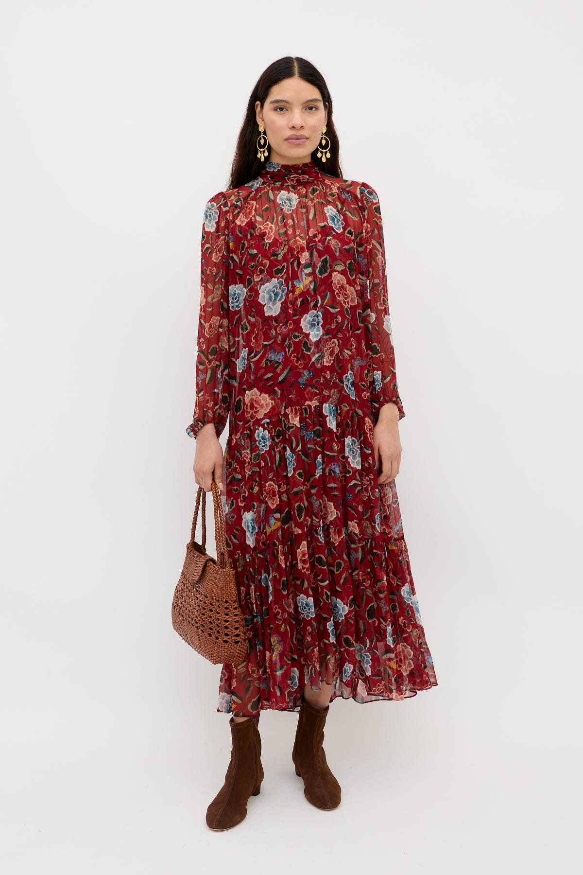 A woman wearing the Adalind - Butterfly Floral Burgundy dress with brown boots, gold earrings, and a brown woven handbag stands against a plain white background.