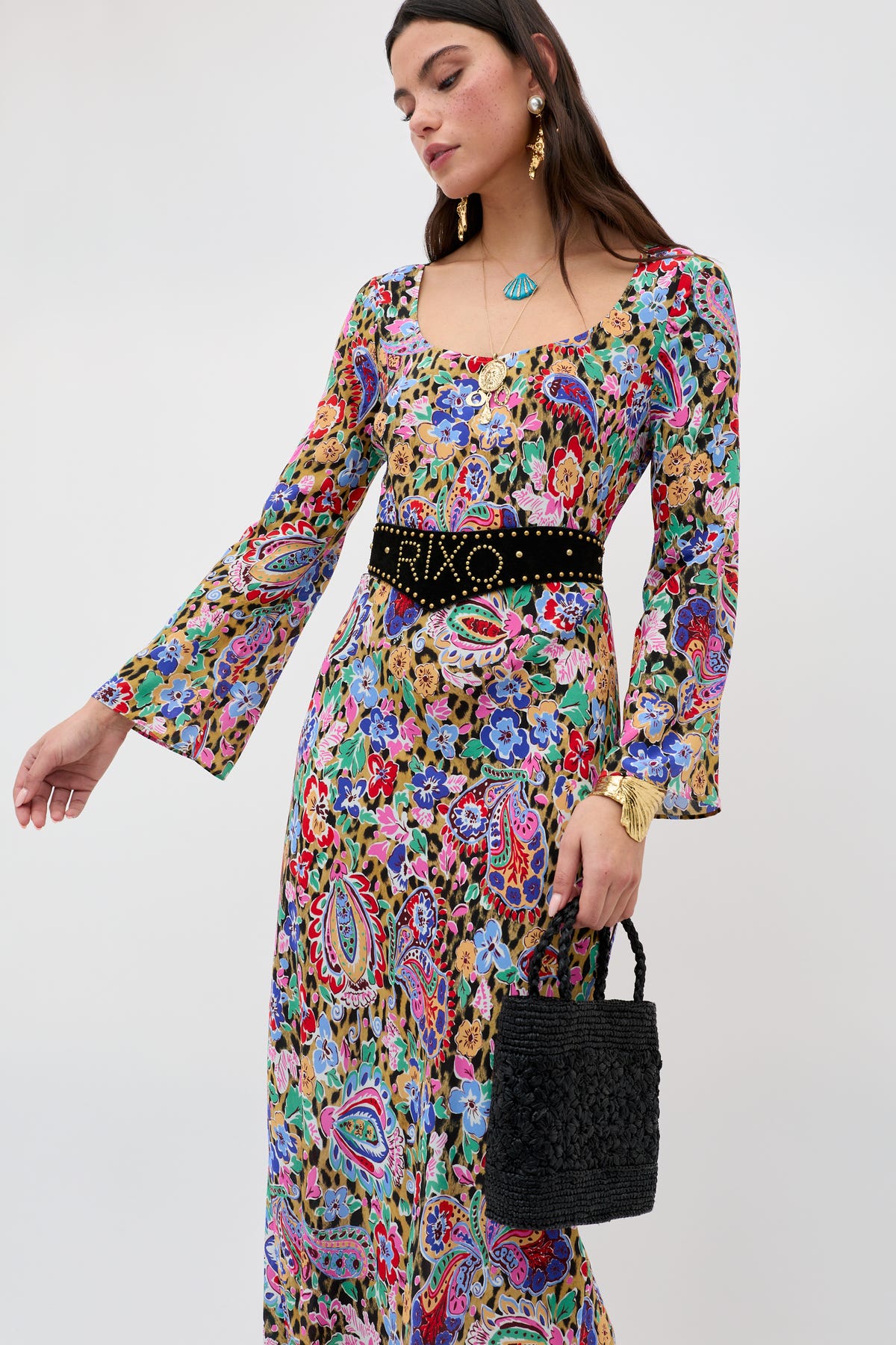 Woman wearing the Noraya - Cara Floral Leopard Brown dress with flared sleeves, styled with a black studded belt, gold earrings, and a turquoise necklace, holding a black woven handbag against a plain background.