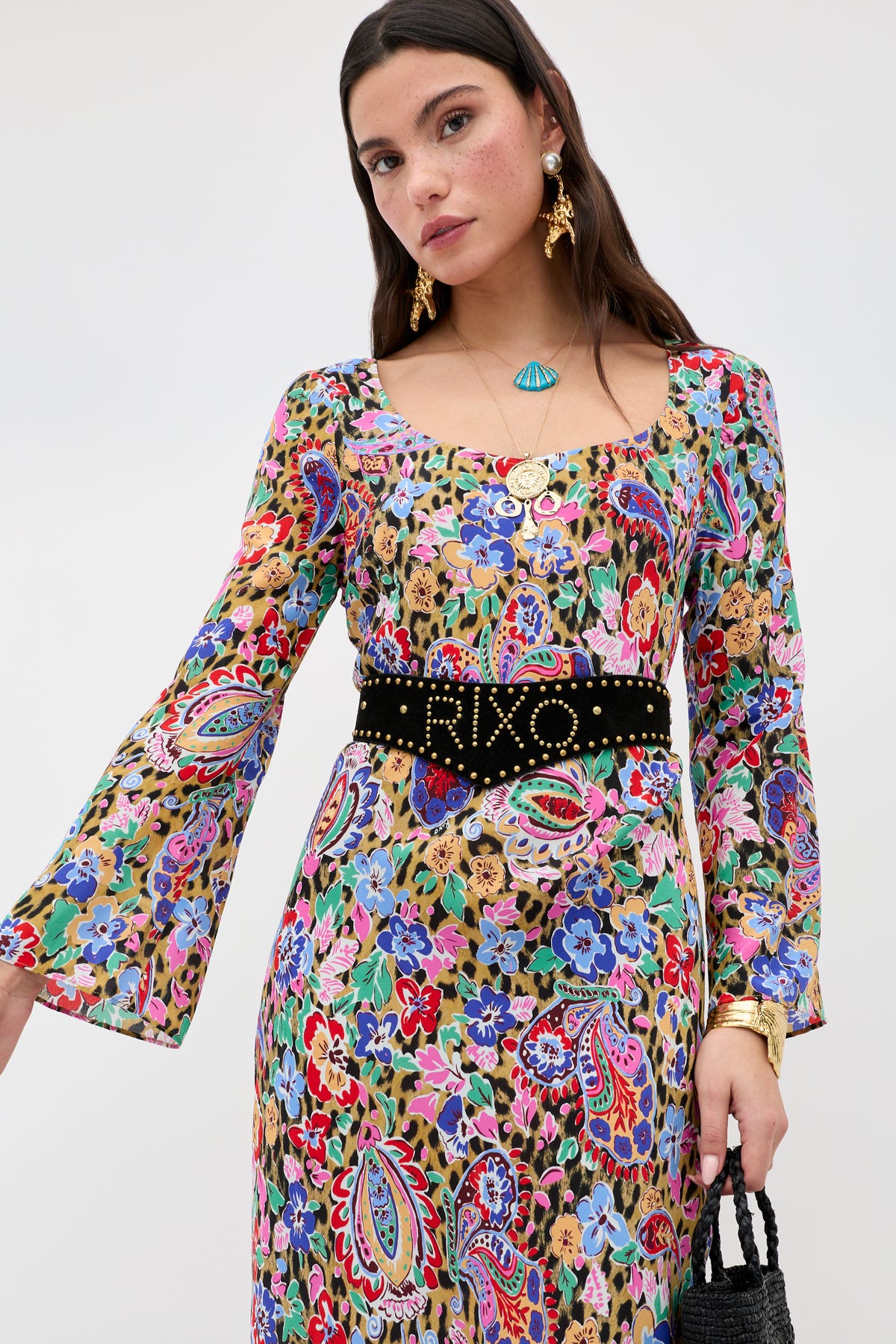 A woman wearing the Noraya - Cara Floral Leopard Brown dress, paired with a black studded belt, gold earrings, a blue pendant necklace, and holding a black bag against a plain background.