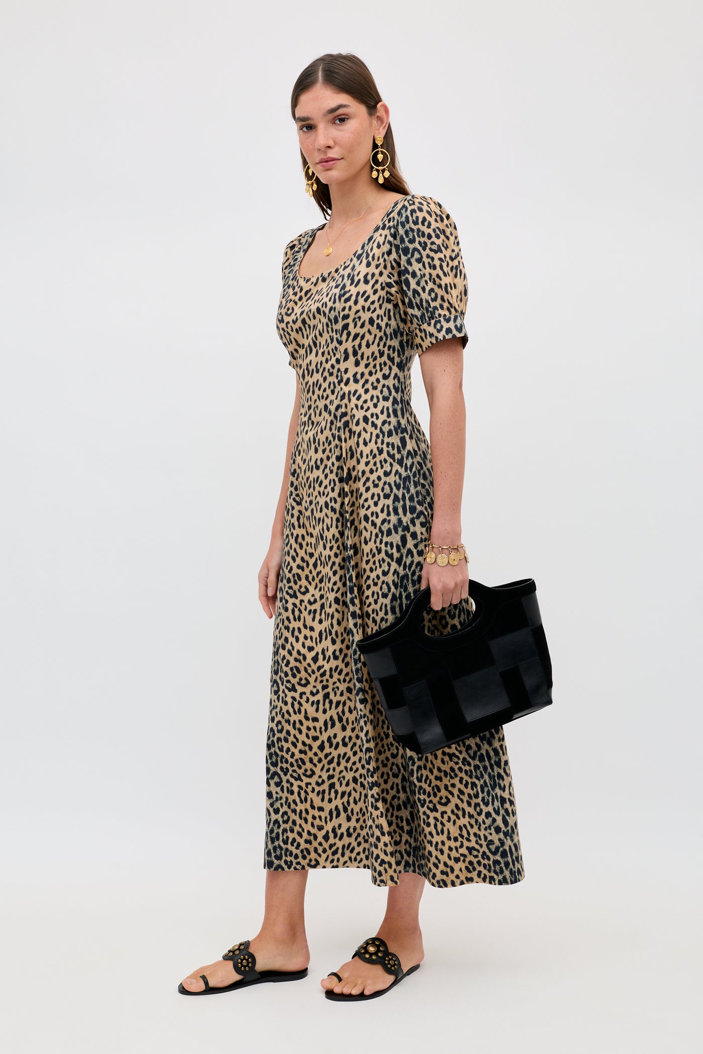 A woman wears the Kathleen - Nina Leopard Brown ankle-length, short-sleeve dress with black sandals and gold jewelry, holding a black handbag against a plain white background.