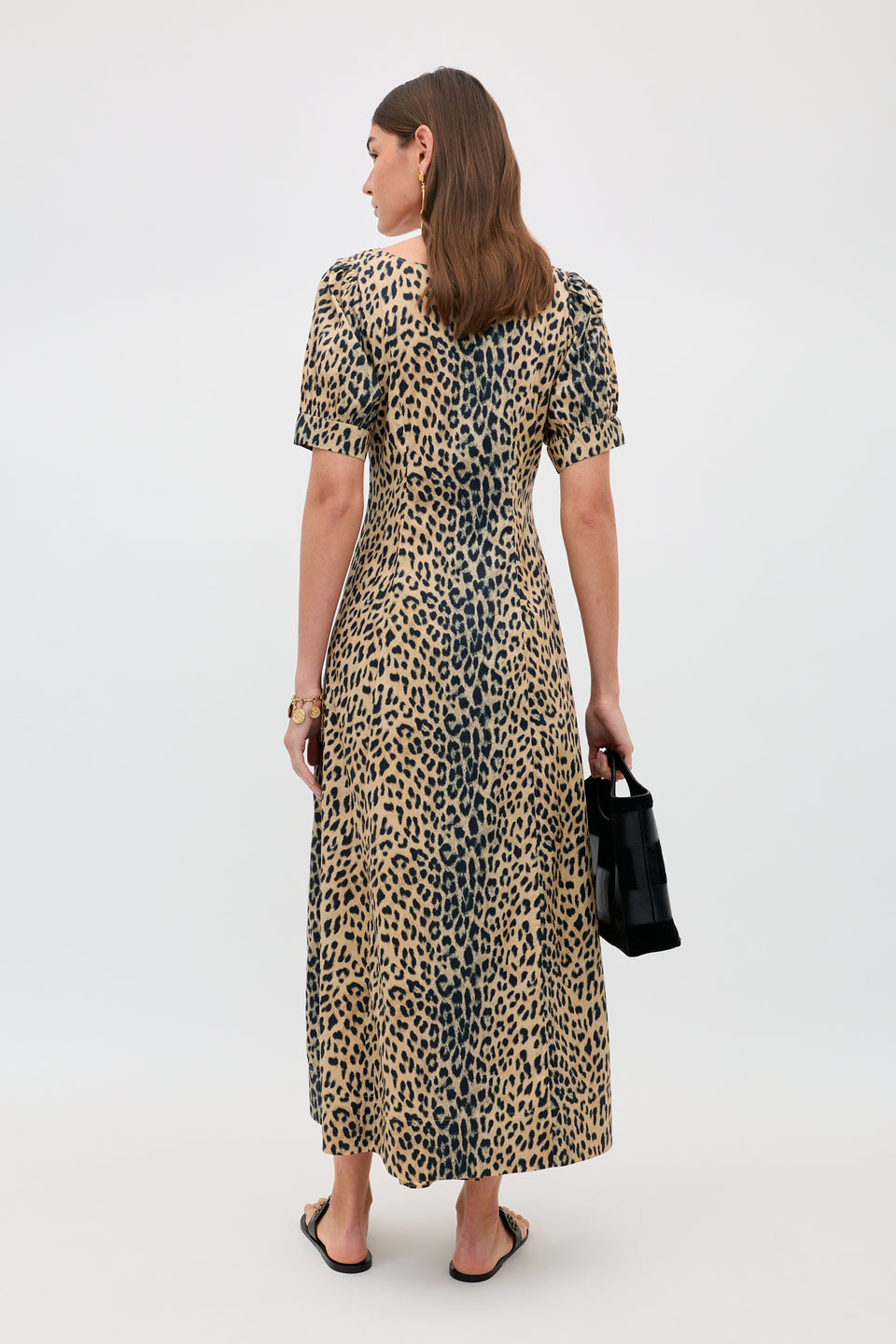 A woman faces away, wearing the Kathleen - Nina Leopard Brown ankle-length dress with short sleeves. She holds a small black purse, wears flat sandals, and her brown hair is loose against a plain light gray background.