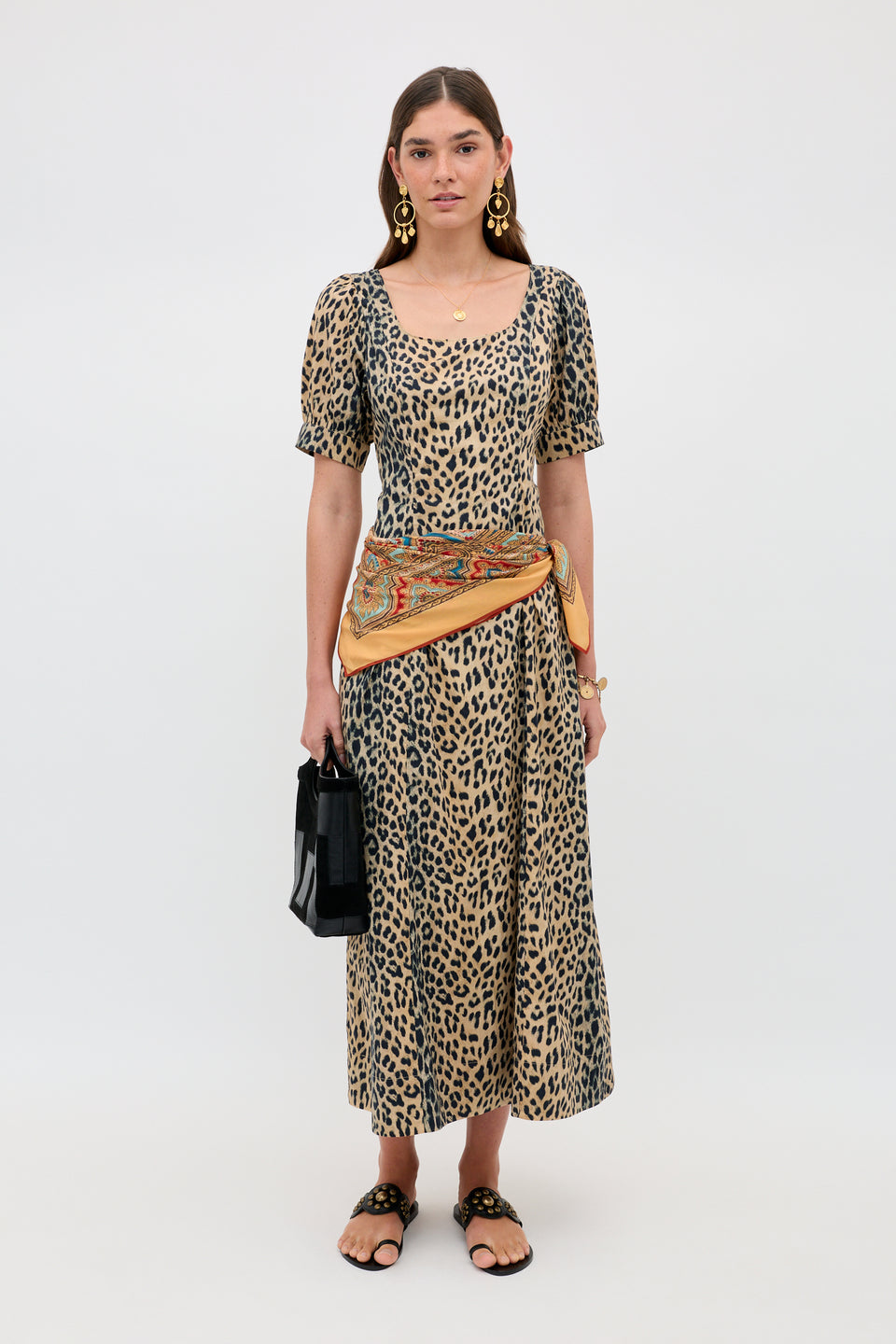 Wearing the Kathleen - Nina Leopard Brown dress, a woman stands against a plain backdrop, styled with statement earrings, sandals, a patterned waist scarf, and a black handbag.