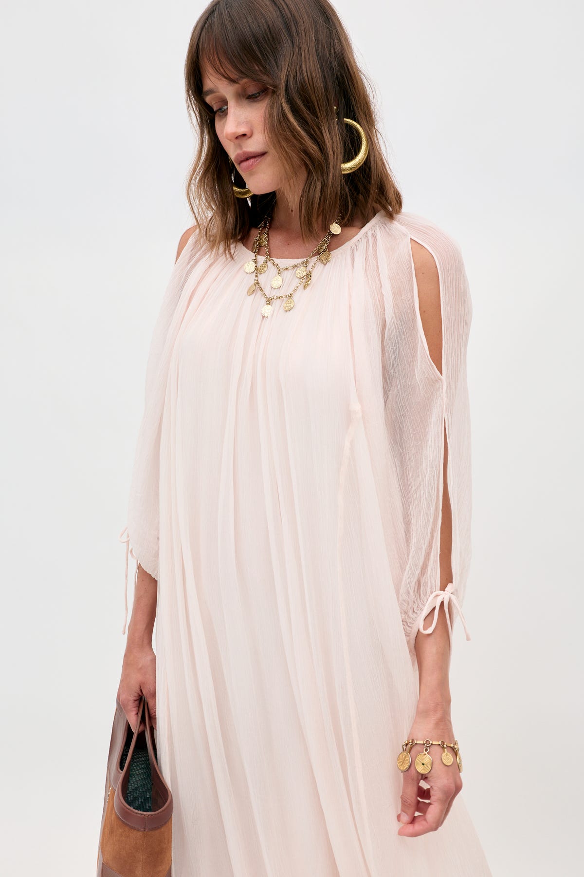 A woman wearing the Hayden - Shell light, flowy blush pink dress with shoulder cutouts, styled with gold hoop earrings, a layered necklace, and bracelet, holds a brown clutch against a plain background.