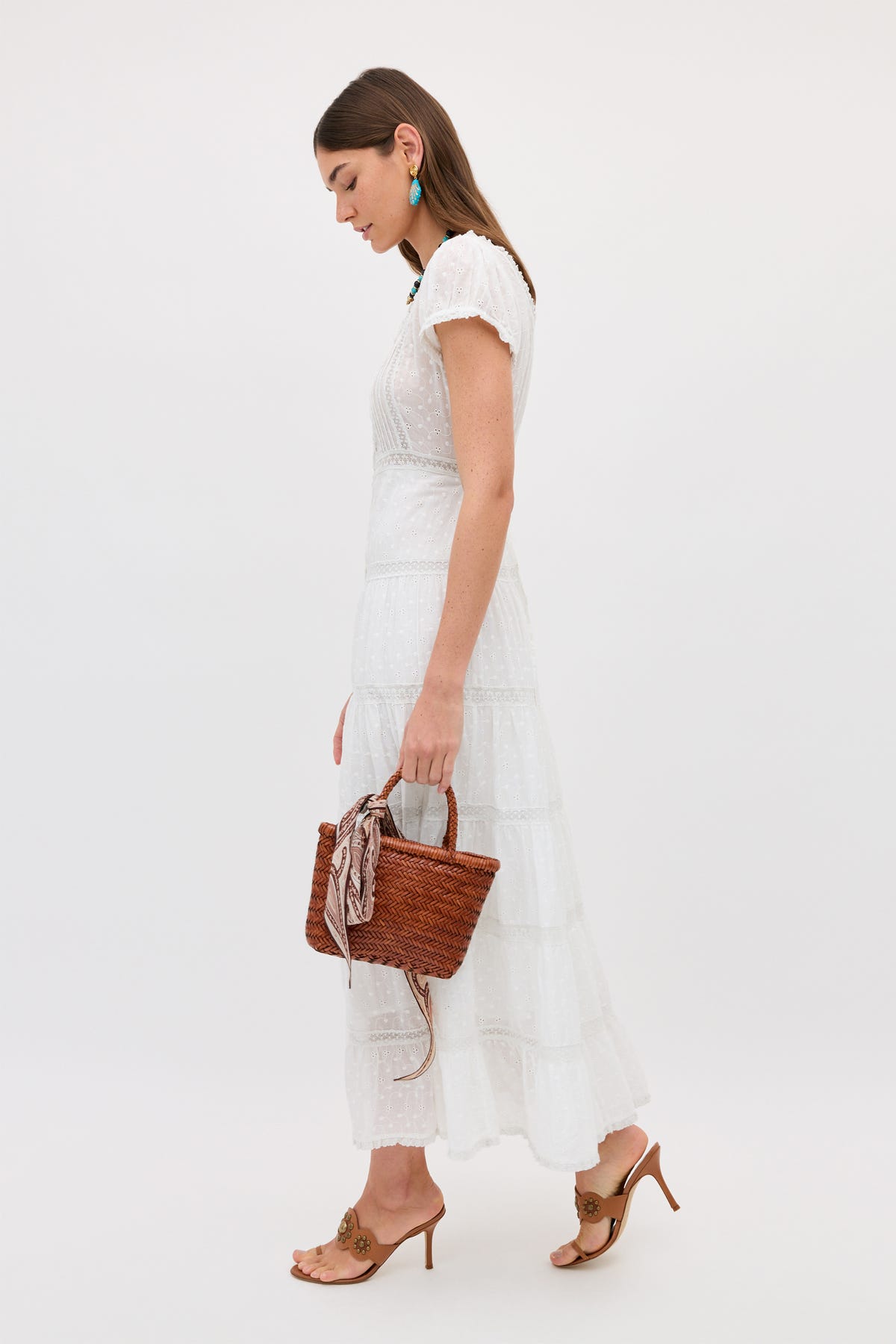 A woman wears the Jaclyn - Ivory cotton summer dress, holding a woven brown handbag with a scarf on the handle. She pairs it with tan heeled sandals and turquoise earrings, standing against a simple light background.