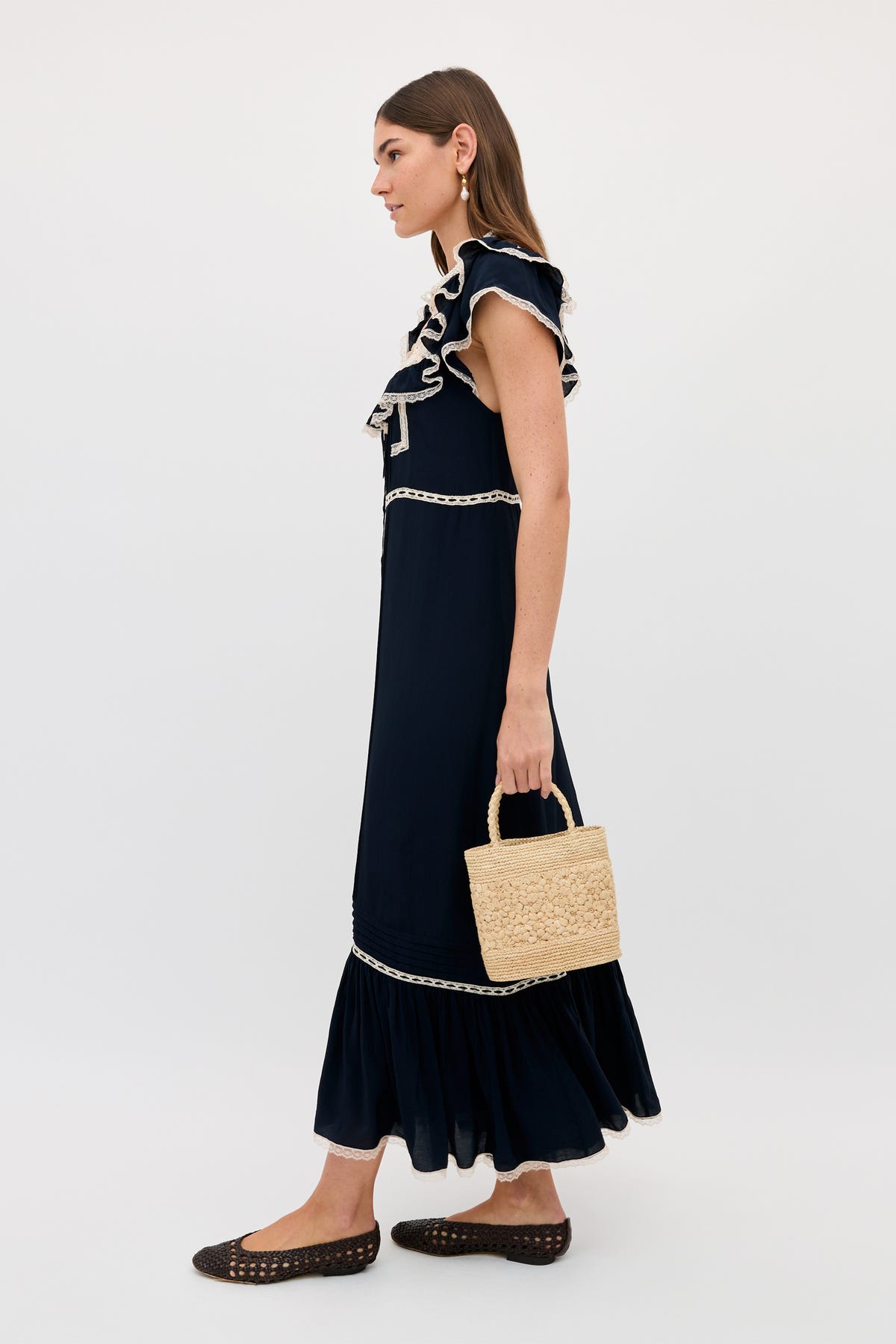 A woman stands in profile wearing the Kendara - Deep Navy cotton midi dress with ruffled details and white trim, holding a woven straw handbag. She pairs it with black woven flats and drop earrings against a plain light background.
