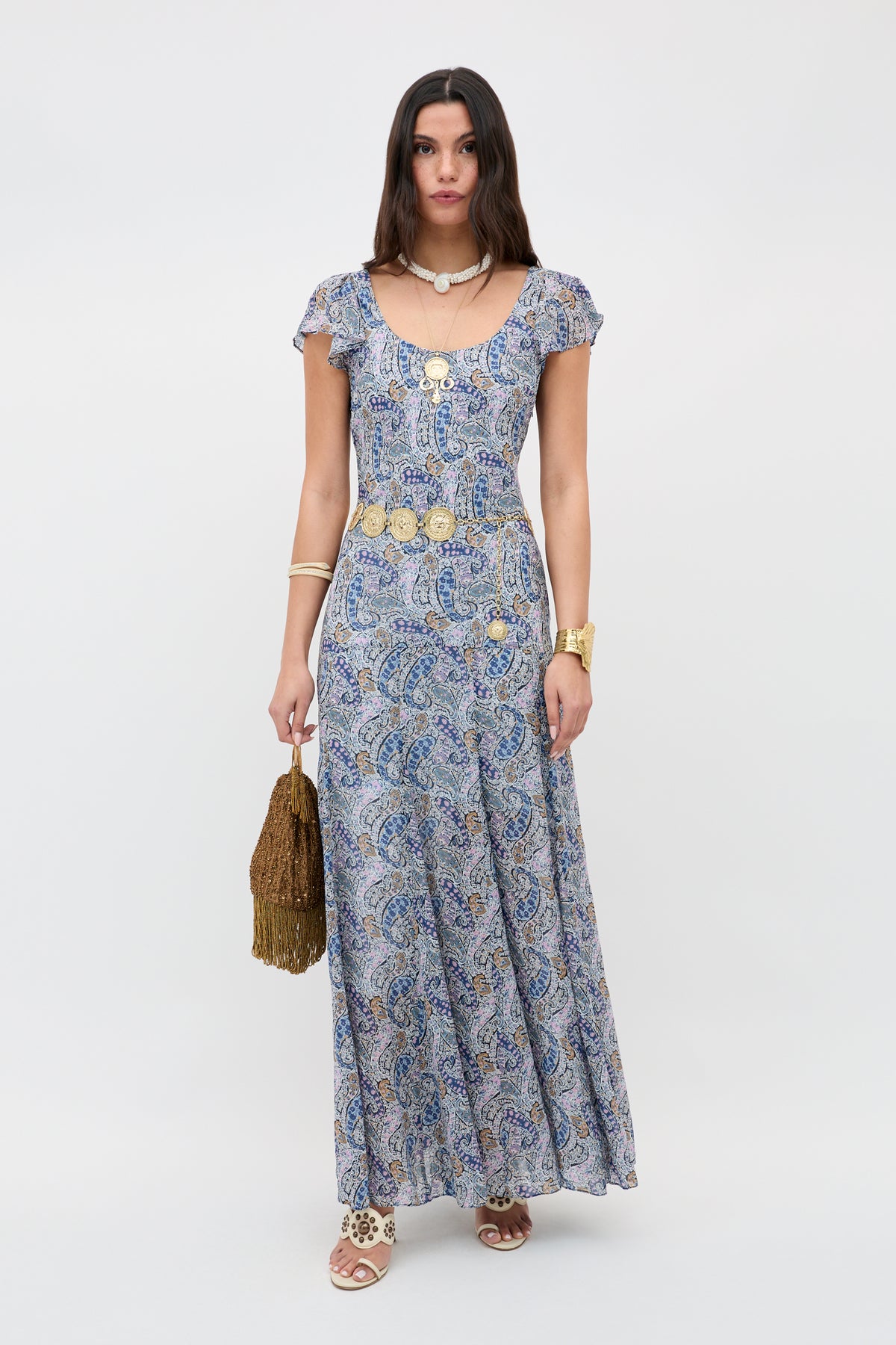 A woman with long straight brown hair stands against a plain background wearing the Erianne - Bourton Navy maxi dress, gold jewelry, sandals, and holding a brown fringed bag.