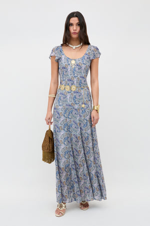 A woman with long straight brown hair stands against a plain background wearing the Erianne - Bourton Navy maxi dress, gold jewelry, sandals, and holding a brown fringed bag.