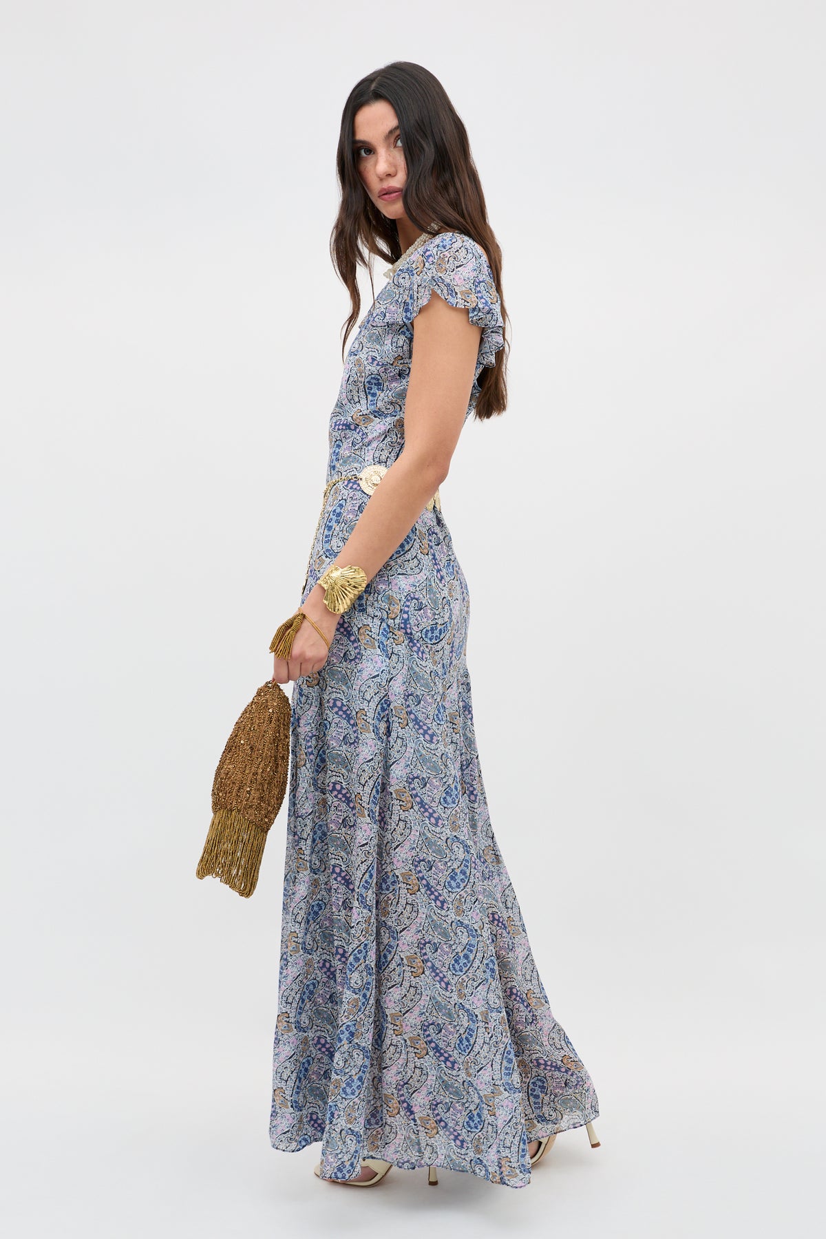 A woman with long dark hair stands sideways against a white background wearing the Erianne - Bourton Navy, a flowy short-sleeved georgette dress in blue and white paisley, paired with heeled shoes, multiple bracelets, and a gold mesh handbag.