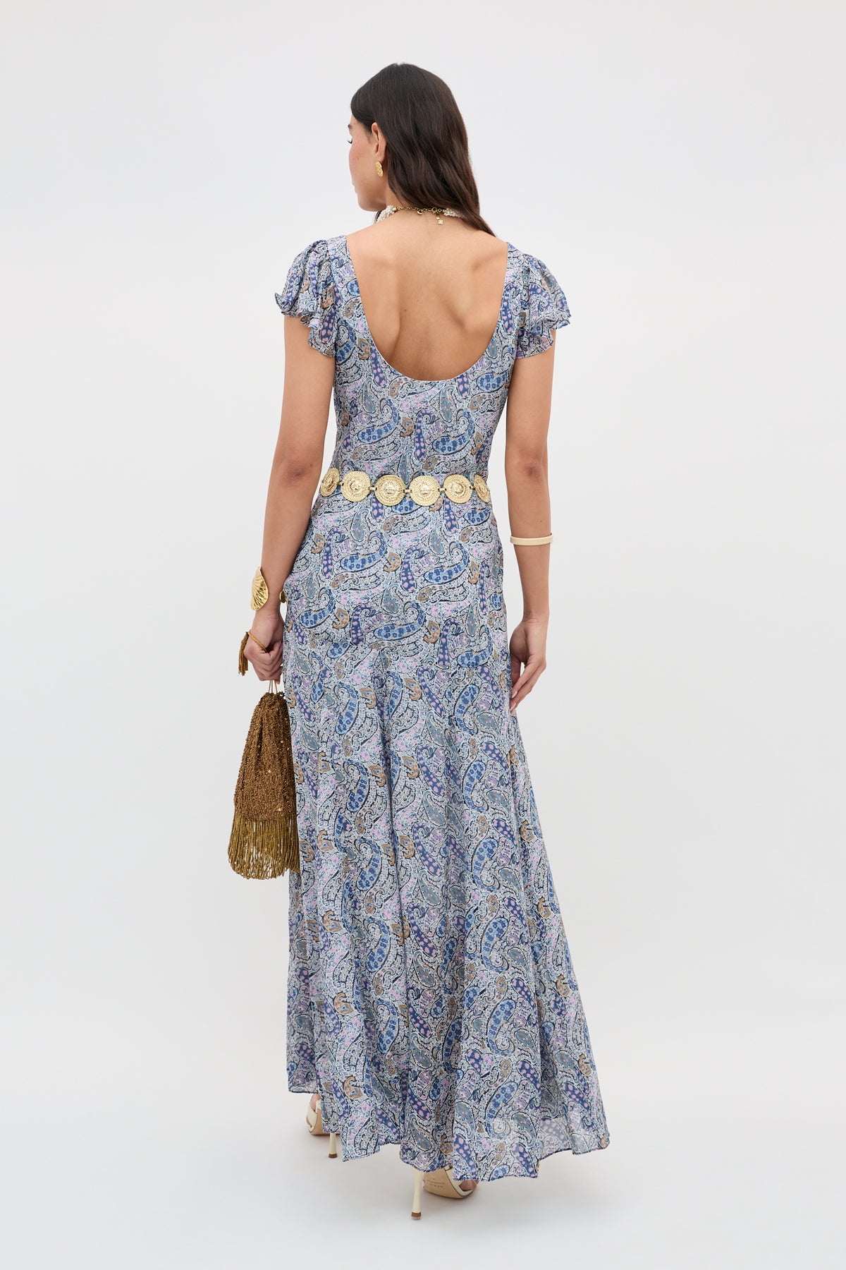 A woman faces away, wearing the Erianne - Bourton Navy dress in blue and white Liberty fabric with a paisley print. She accessorizes with a gold fringed bag, decorative belt, and bracelets.